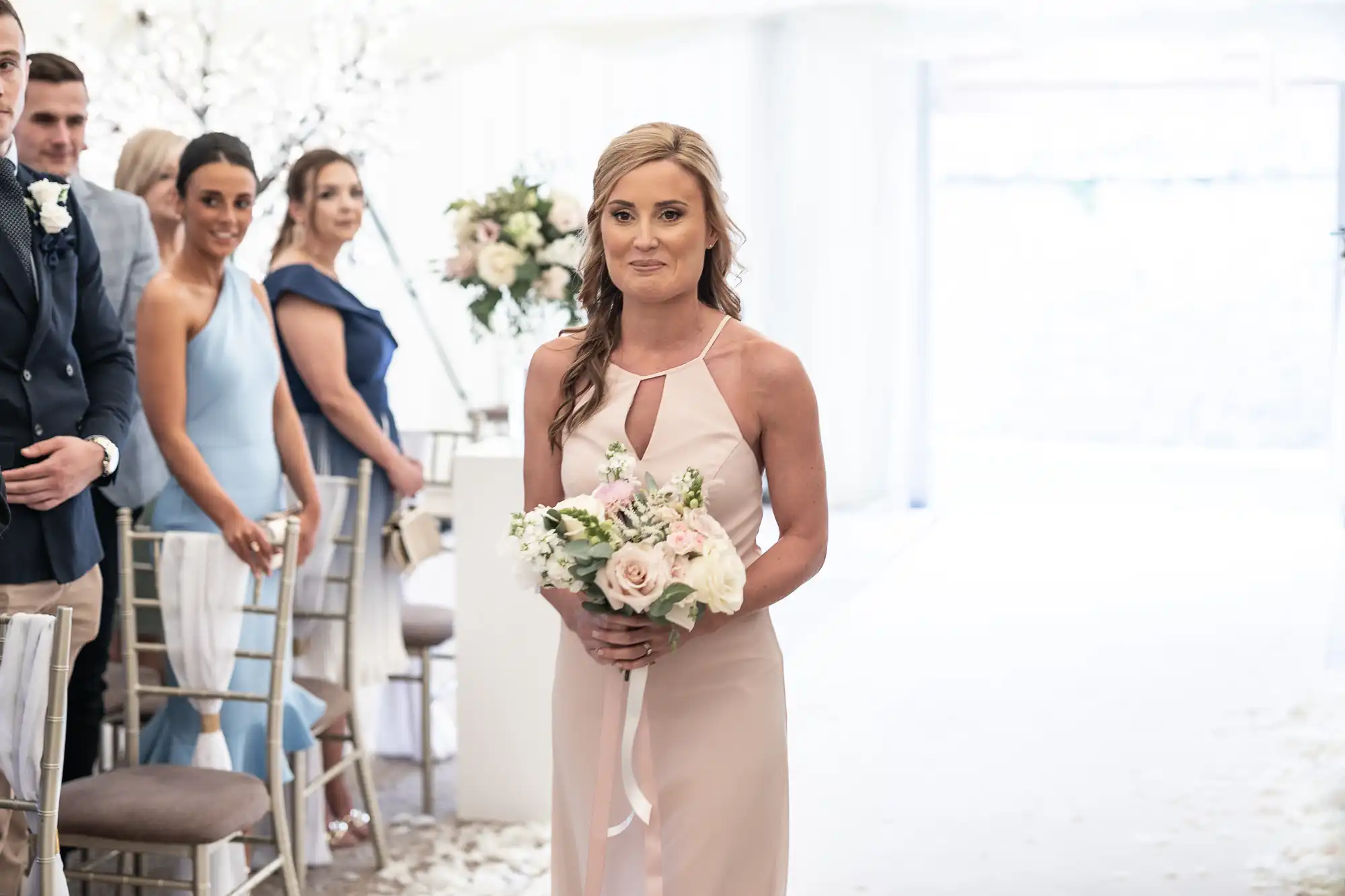 A woman in a light pink dress holds a bouquet of flowers while walking down an aisle. People in formal attire are seated and standing on either side of the aisle.