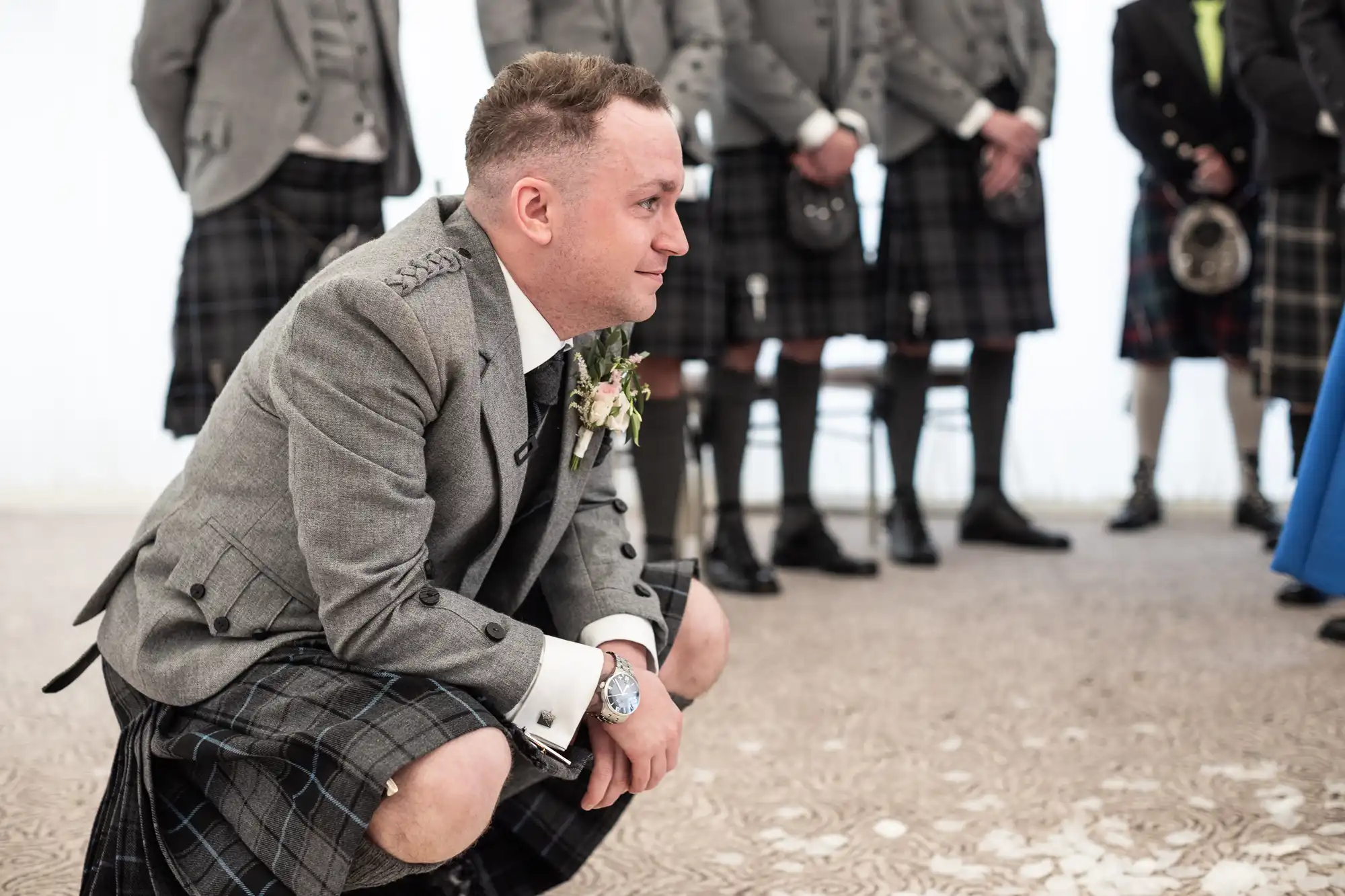 A man in a gray suit jacket and kilt crouches on the ground with a serious expression, surrounded by other men in similar attire.