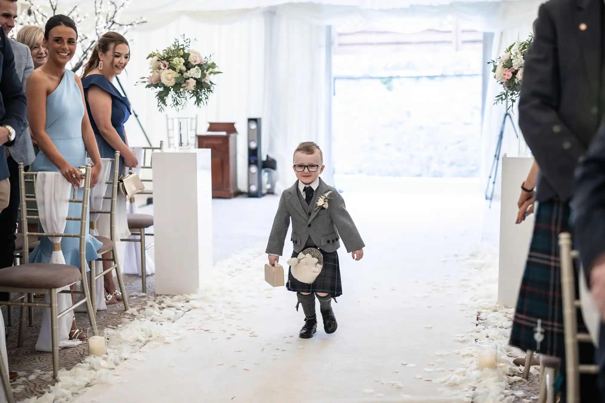 A young boy dressed in a grey suit and kilt walks down a flower-petal-strewn aisle at a wedding ceremony, holding a ring bearer pillow. Guests are seated on either side, watching and smiling.