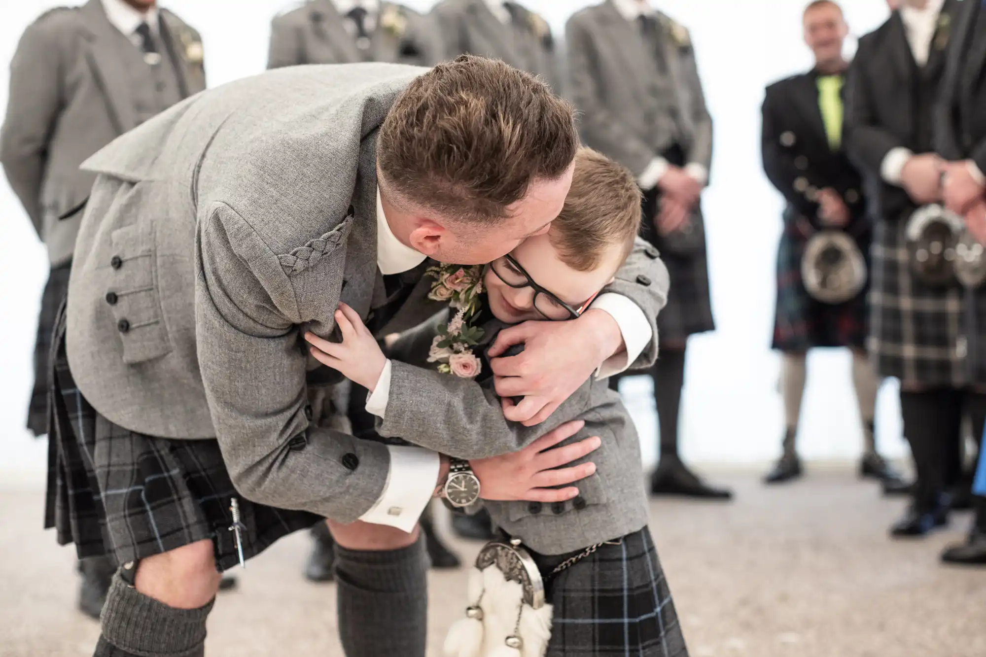 An adult in a grey suit and kilt kneels down to hug a child in a matching outfit among a group of similarly dressed men.