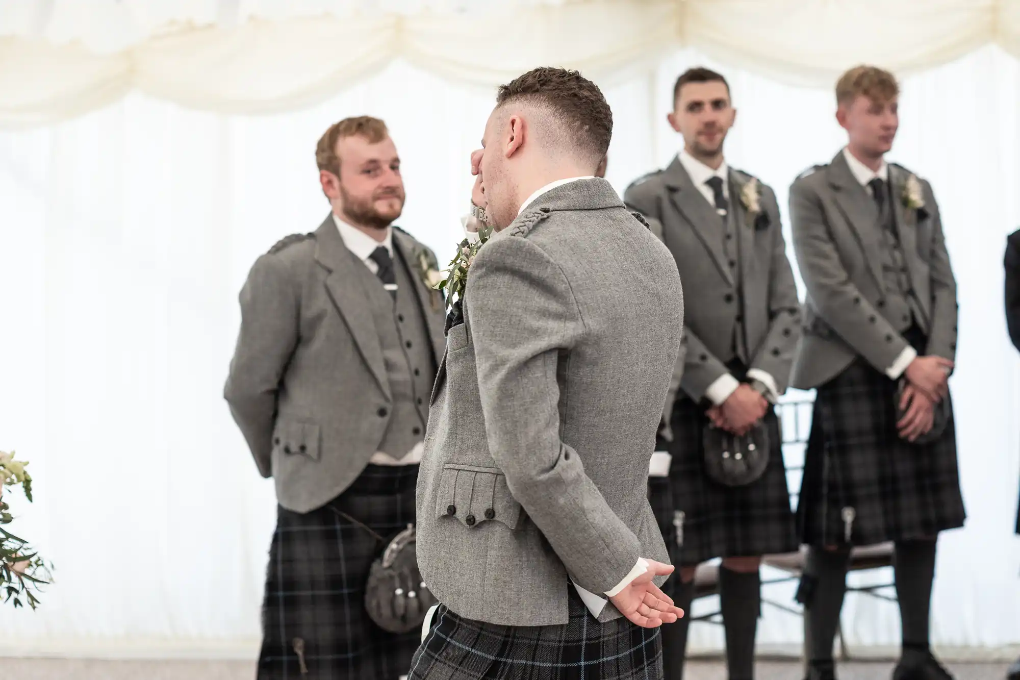 Several men in grey formal attire, including tartan kilts, stand in a line, with one man facing away and holding his hands behind his back. They appear to be at a formal event.