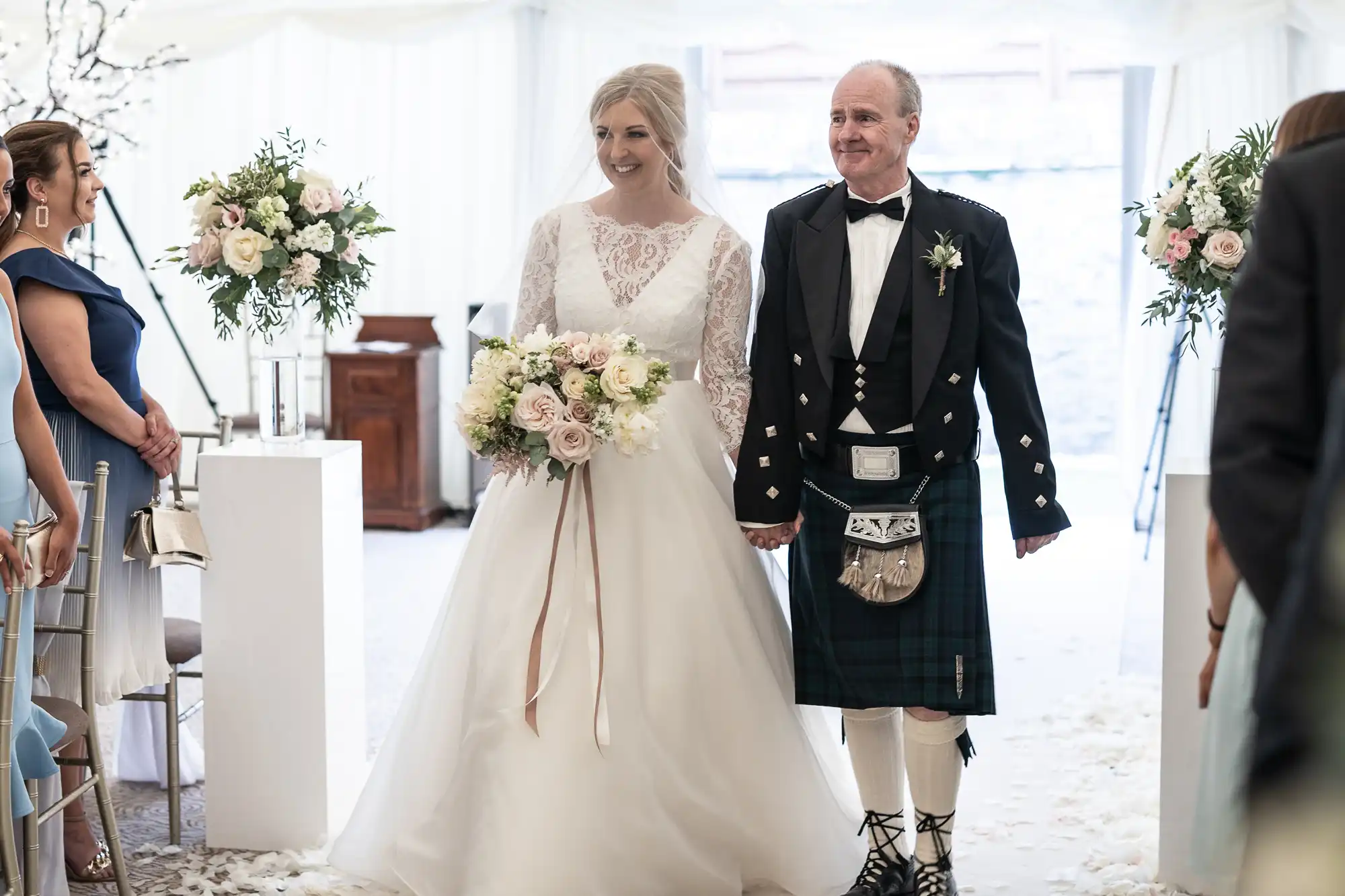 A bride in a white lace wedding dress walks down the aisle holding hands with a man in a traditional Scottish kilt. They are smiling and surrounded by guests and floral arrangements.