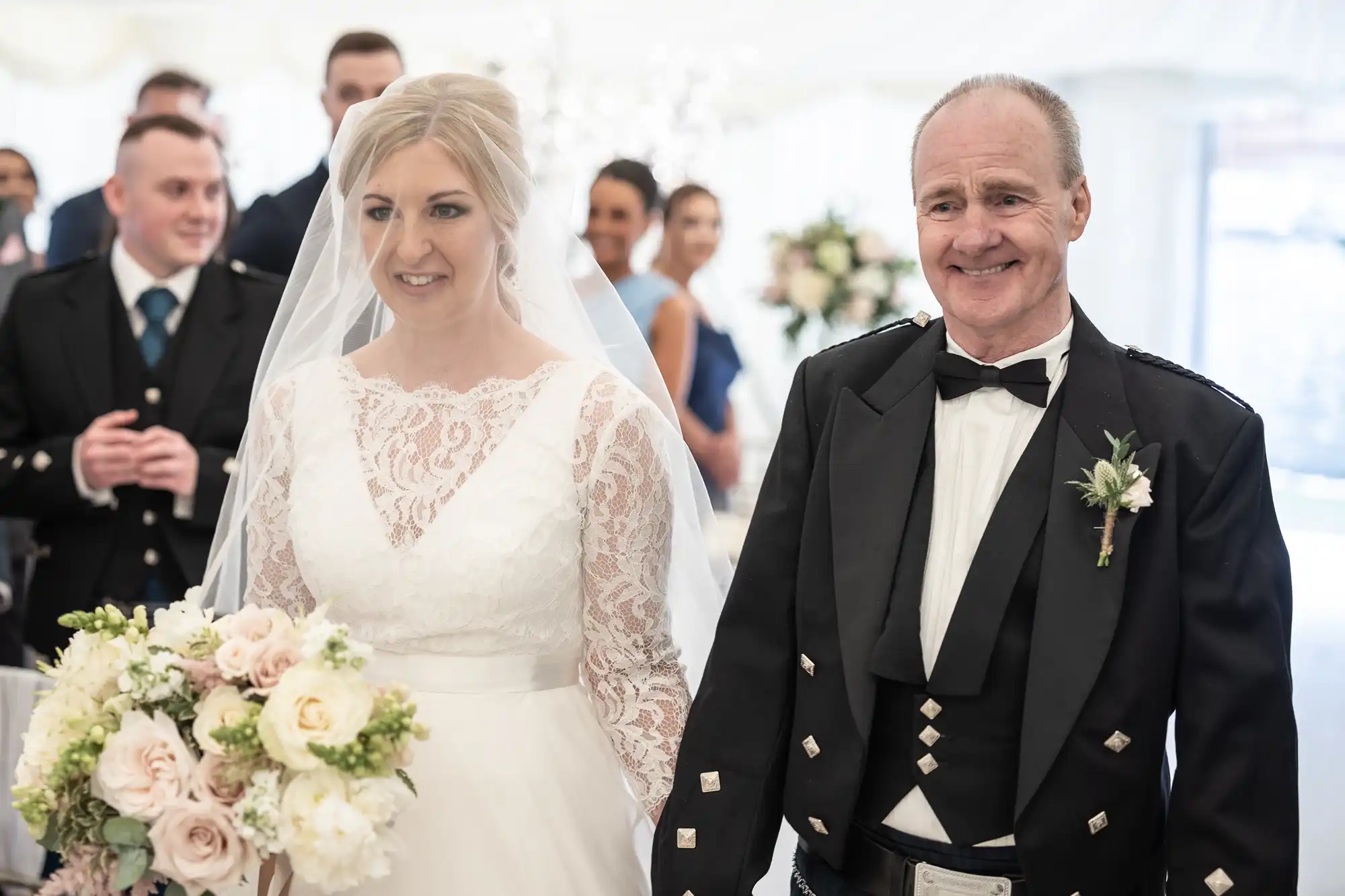 A bride in a white lace dress stands with an older man in formal attire. She holds a bouquet of flowers, and several people in formal wear are visible in the background.