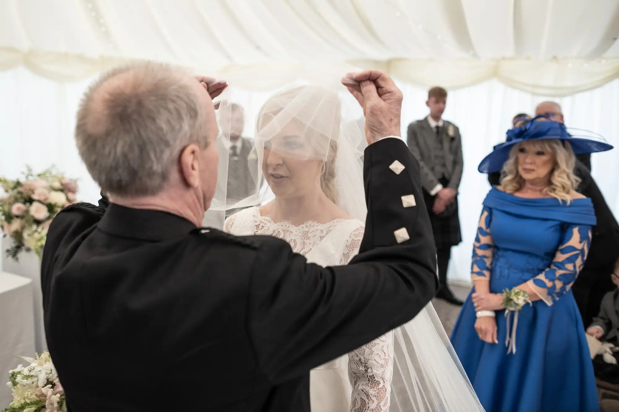 An older man lifts the veil of a bride in a white dress during a wedding ceremony. The bride smiles, and guests, including a woman in a blue dress and hat, watch in the background.