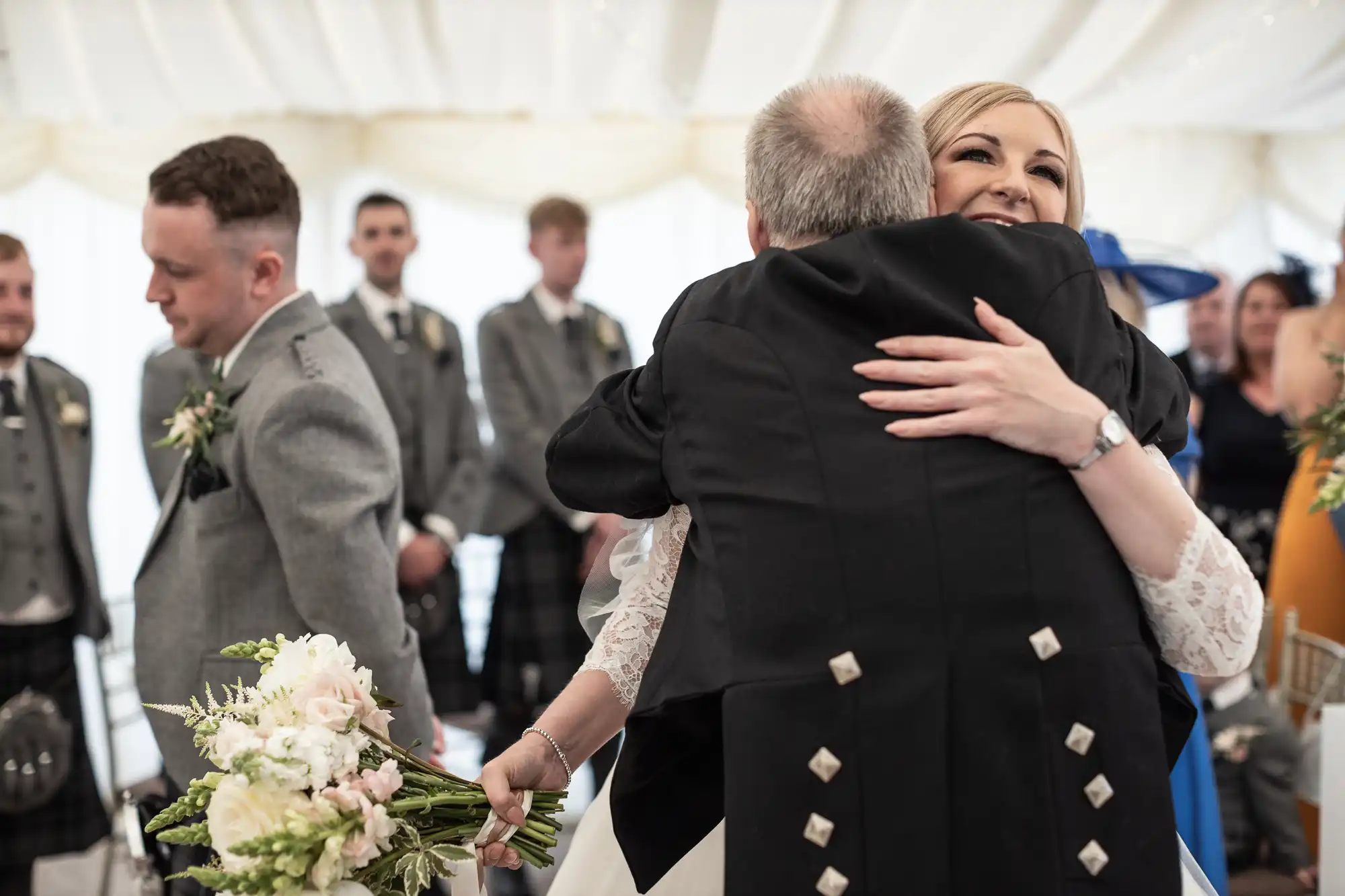 A bride hugs an older man in a suit while holding a bouquet. Several men in gray attire stand in the background.