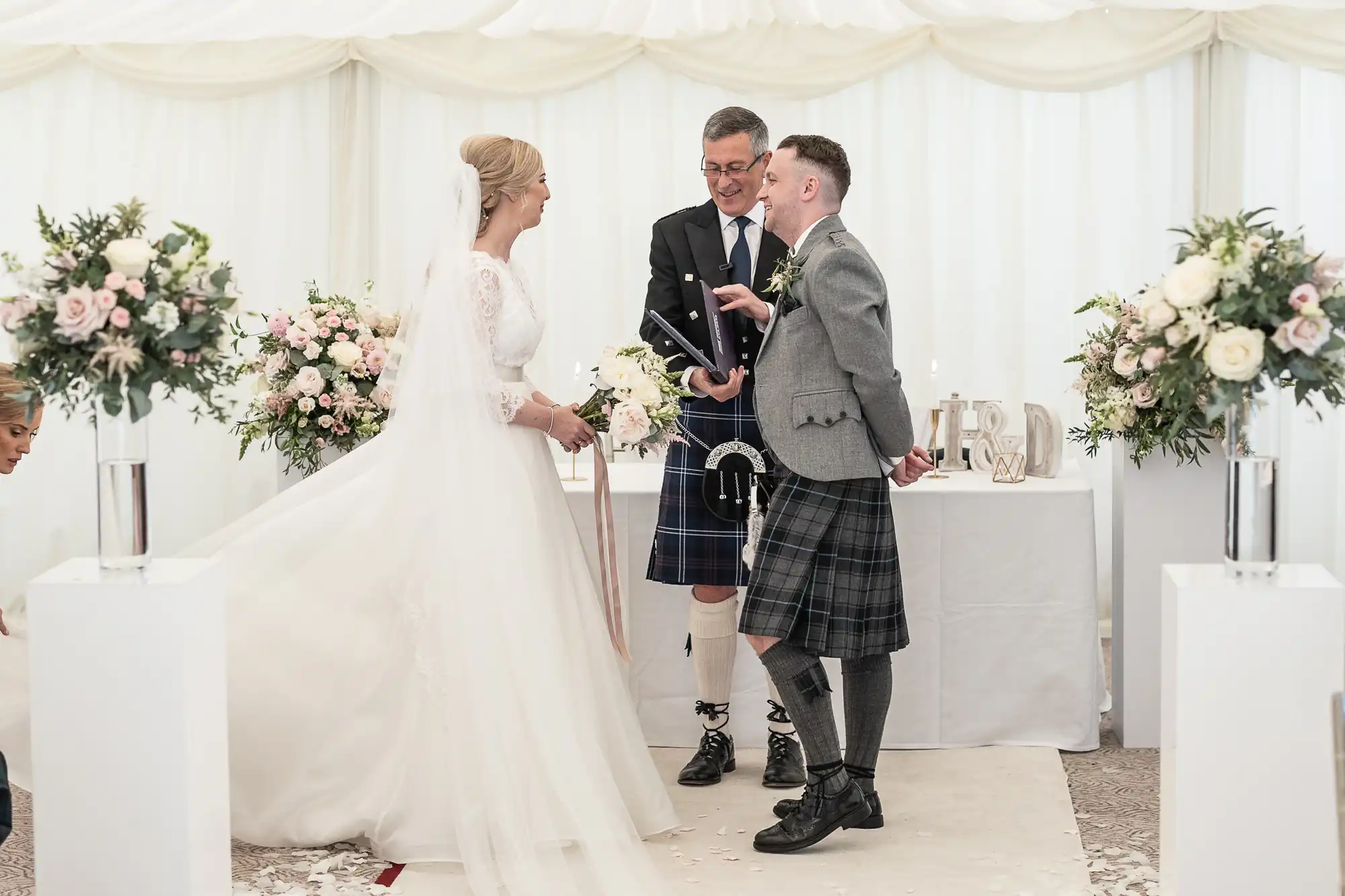 A bride and groom, both wearing traditional Scottish attire, are standing at the altar with the officiant in a decorated white tent. The groom is holding a book and smiling.