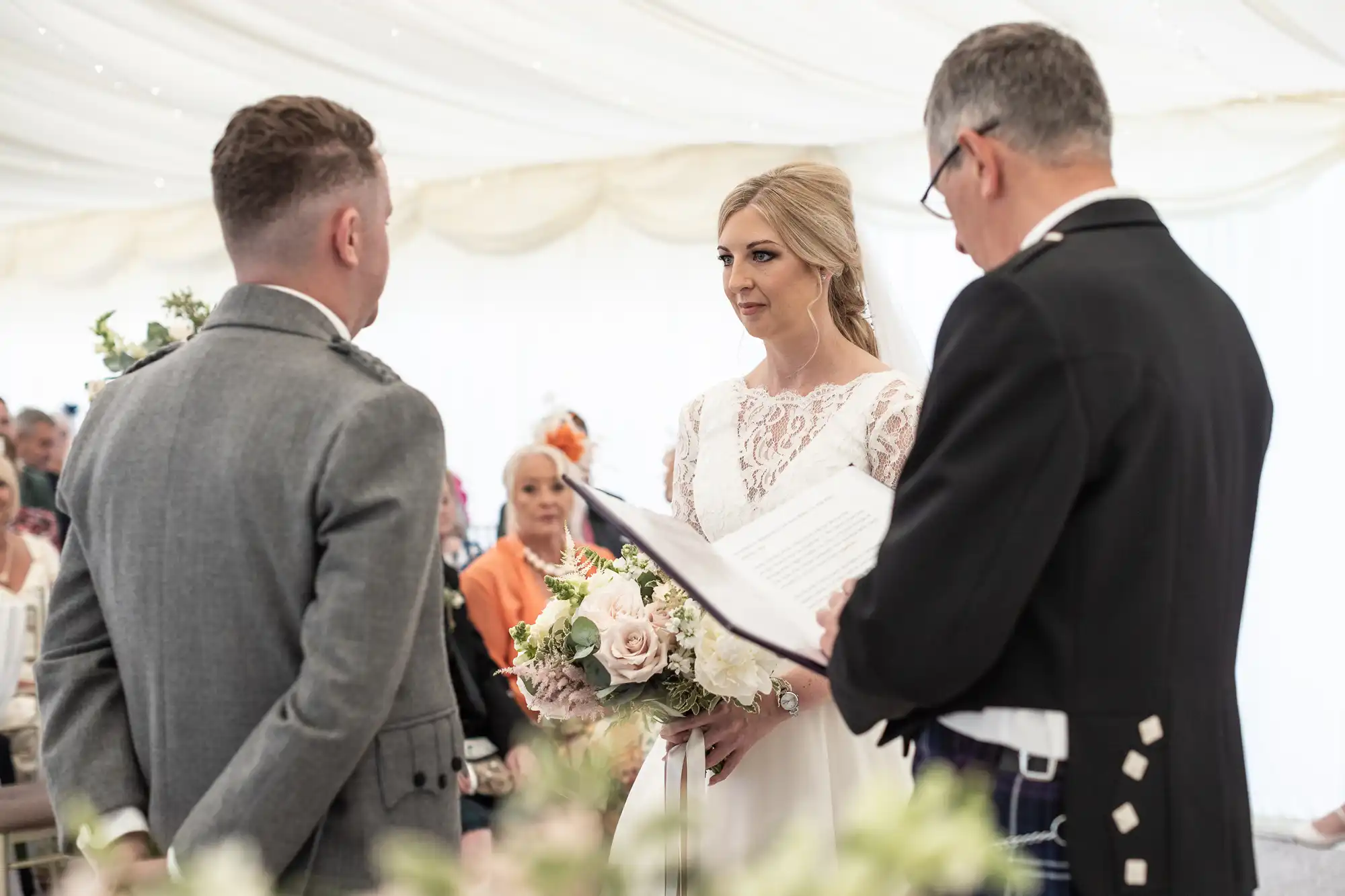 A bride and groom stand facing each other during a wedding ceremony, with the officiant reading from a book. The bride holds a bouquet of flowers.