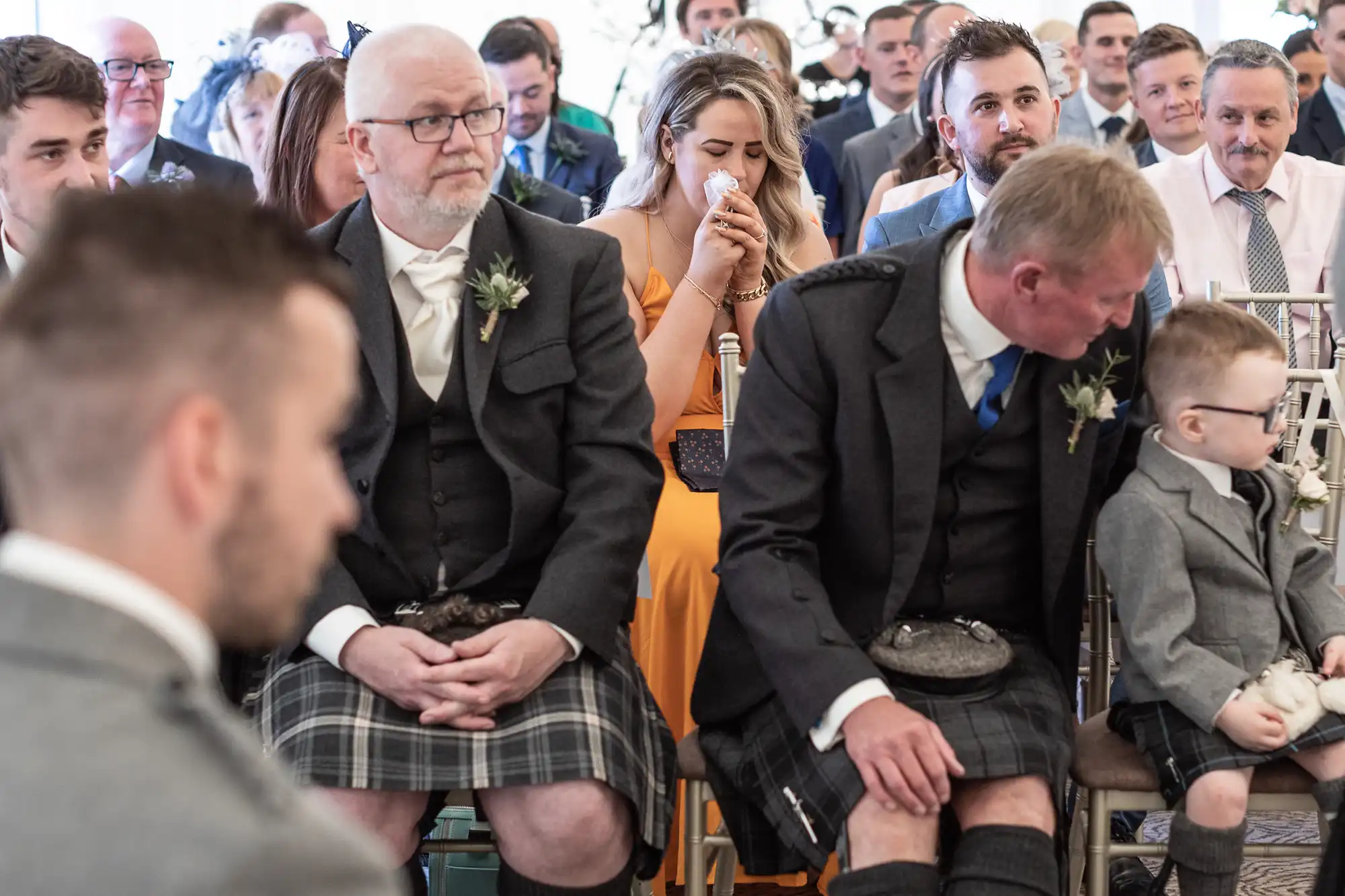 A group of people, many in formal attire, sit closely together at an event. A woman in the center is wiping her nose, and several men are wearing kilts.