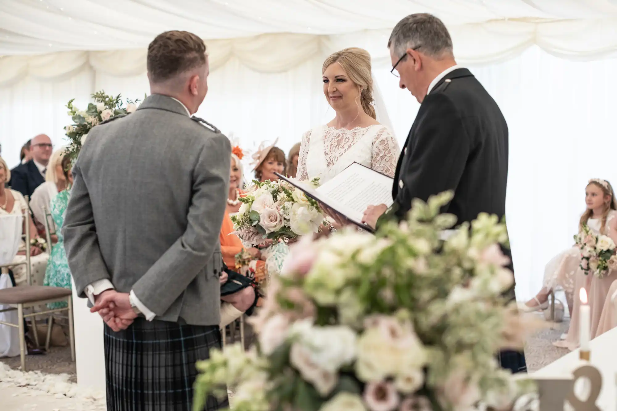 A bride and groom stand before an officiant during a wedding ceremony in a decorated room, with guests seated in the background.