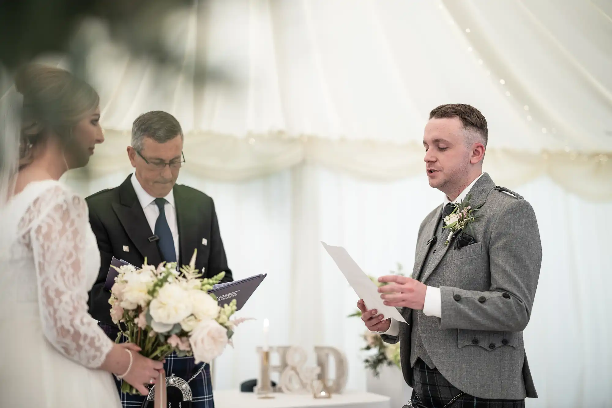 A groom reads from a paper during a wedding ceremony while the bride and officiant listen. The couple stands in front of a table with floral arrangements and decorative initials.