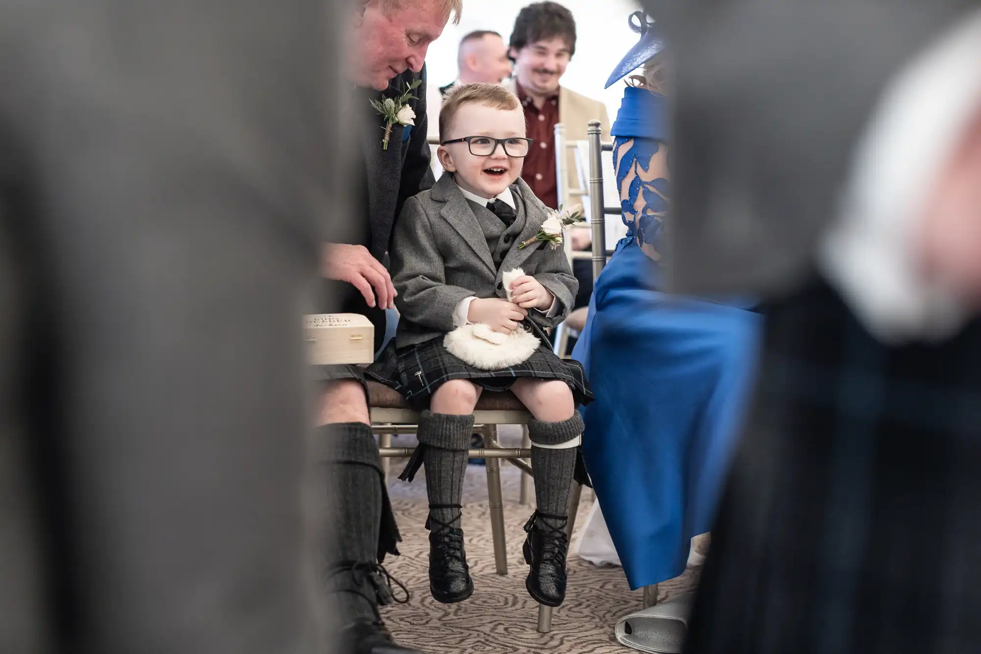 A young boy in a kilt, glasses, and formal grey attire sits on an adult's knee, smiling while holding a flower, with blurred people seated in the background.