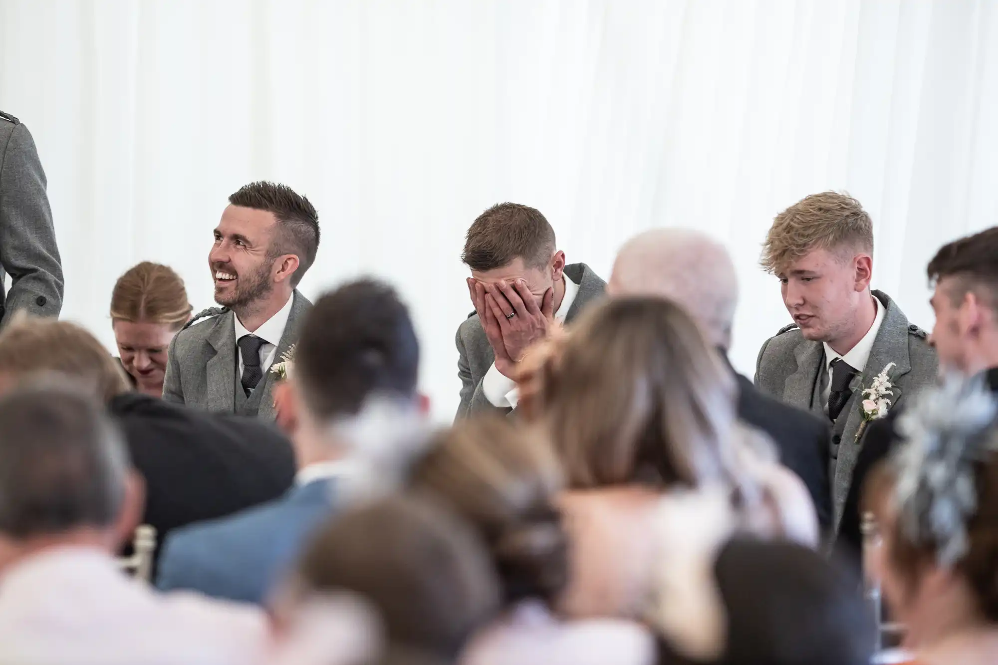 A group of people in formal attire are seated, with one man covering his face with his hands, appearing emotional. Others around him are looking on or smiling.