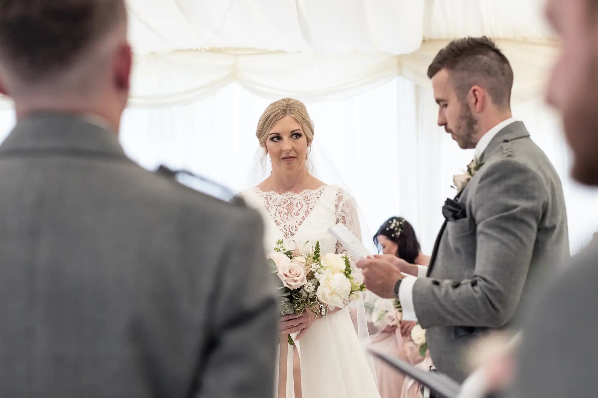 A bride and groom stand in a decorated venue during their wedding ceremony; the groom reads from a piece of paper while the bride holds a bouquet and looks ahead.