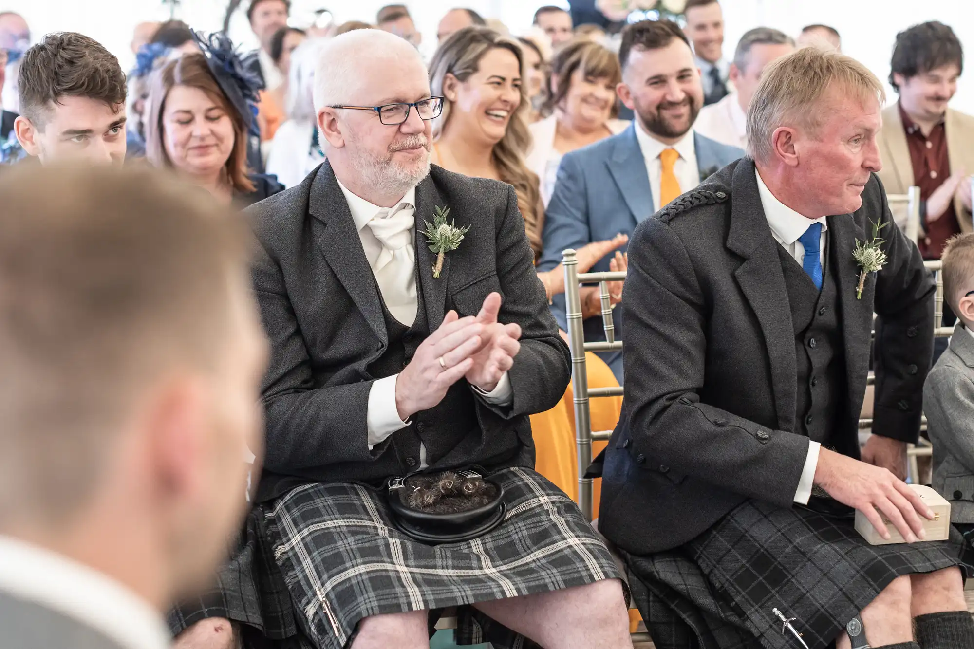 A group of people sits in an indoor setting, with many wearing formal attire. Two men in the foreground wear kilts, with one clapping and the other seated beside him. Others in the background are seen smiling.