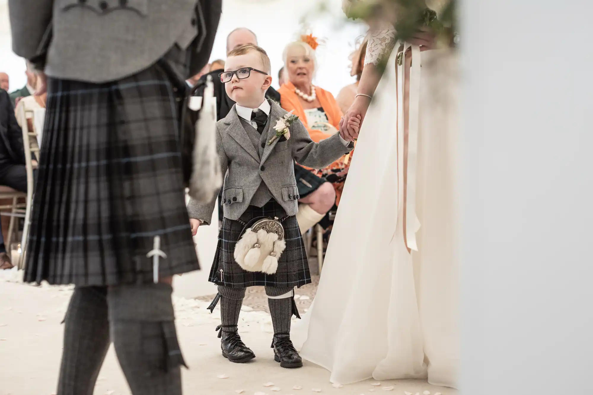 A young boy dressed in traditional Scottish attire stands holding hands with an adult at a wedding ceremony, with seated guests in the background.