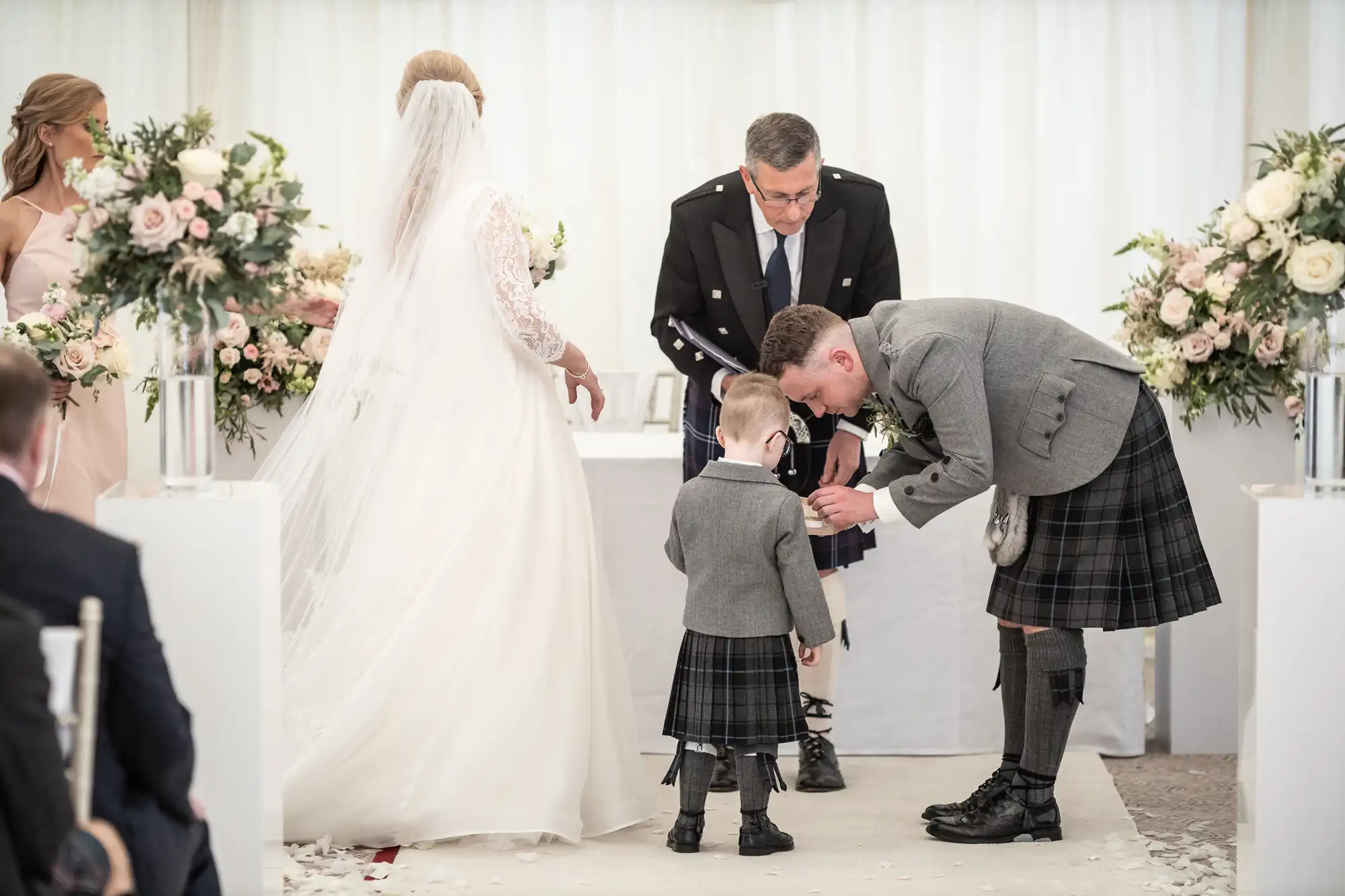 A groom in a kilt bends down to talk to a young boy in matching attire during a wedding ceremony. A bride in a white gown stands nearby, accompanied by other attendees and floral arrangements.