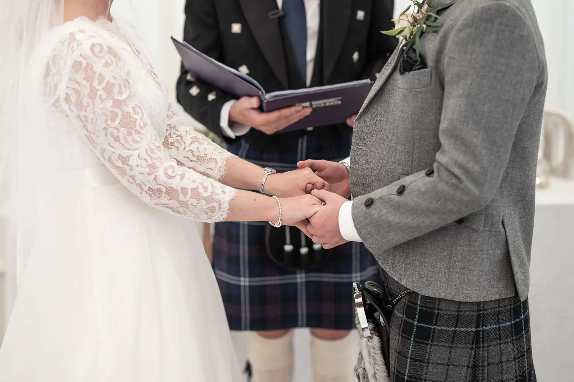 A couple holding hands during their wedding ceremony, both dressed in traditional attire, with an officiant standing between them holding an open book.