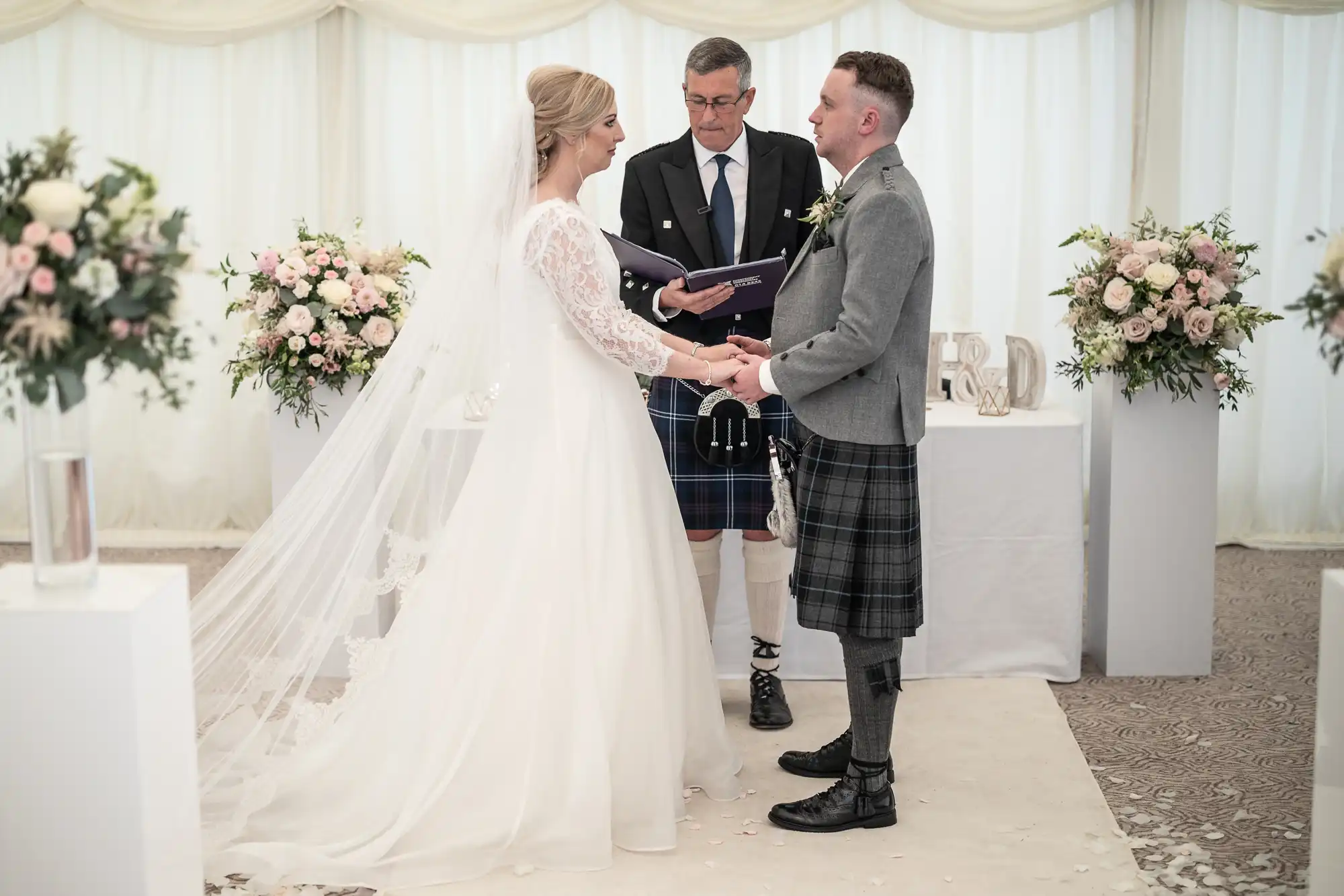 A bride and groom holding hands during their wedding ceremony, with an officiant standing behind them. The groom and officiant wear Scottish kilts, and floral arrangements decorate the background.