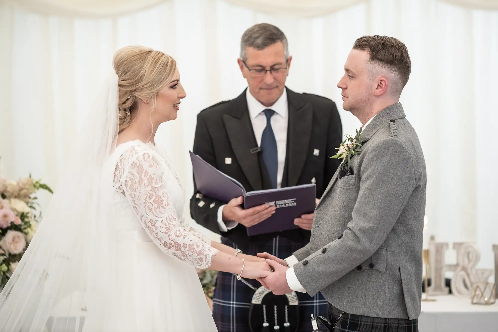 A bride and groom holding hands stand facing each other. An officiant, standing behind them, reads from a book during the wedding ceremony.