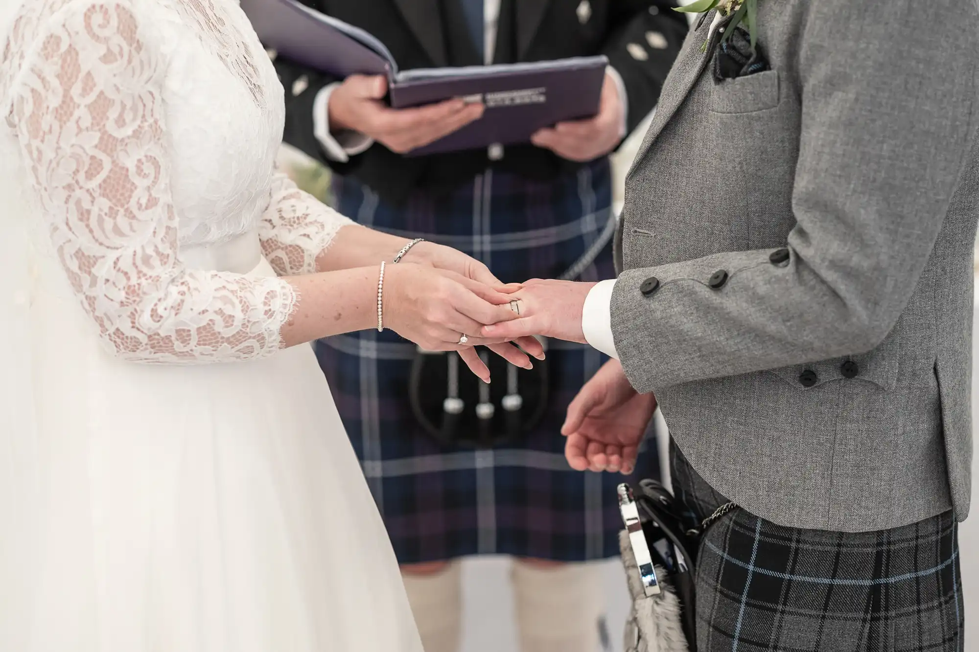 A bride and groom exchange rings during a wedding ceremony. The officiant, dressed in a kilt, holds a book near them. The bride wears a white dress, and the groom wears a grey suit with a kilt.