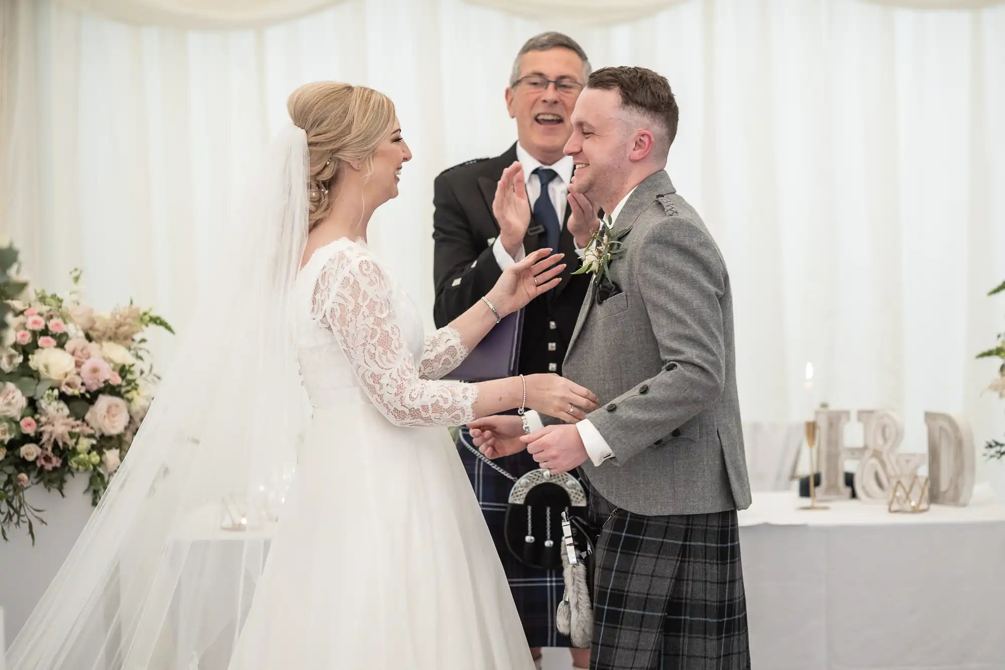 A bride and groom exchange smiles and hold hands while a man behind them applauds. They are in a decorated indoor setting with floral arrangements and a table in the background.