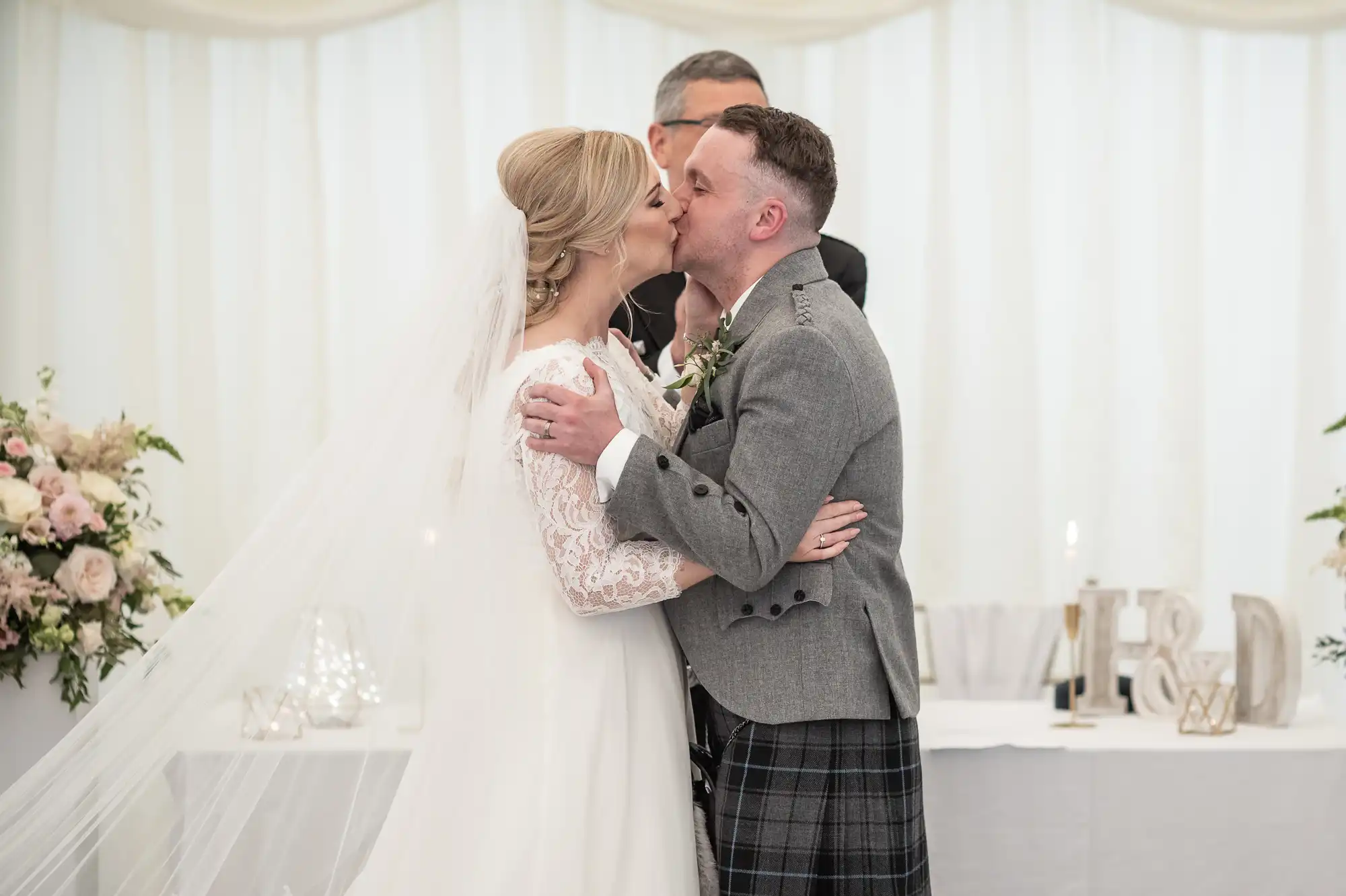 A bride and groom share a kiss at their wedding ceremony. The bride is in a white dress and veil, and the groom is in a gray jacket and kilt. Flowers and decorations are visible in the background.