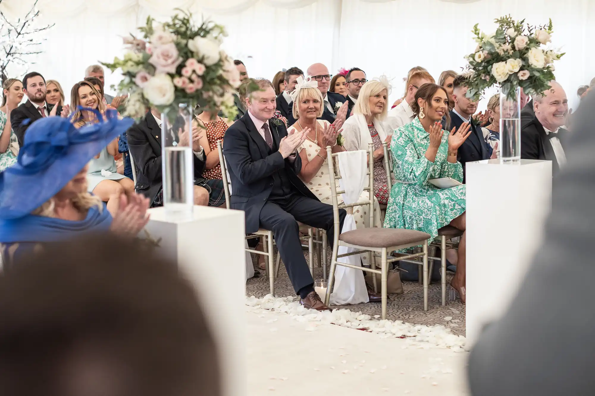 A seated audience in formal attire claps during an indoor event, with flower arrangements on pillars in the foreground.