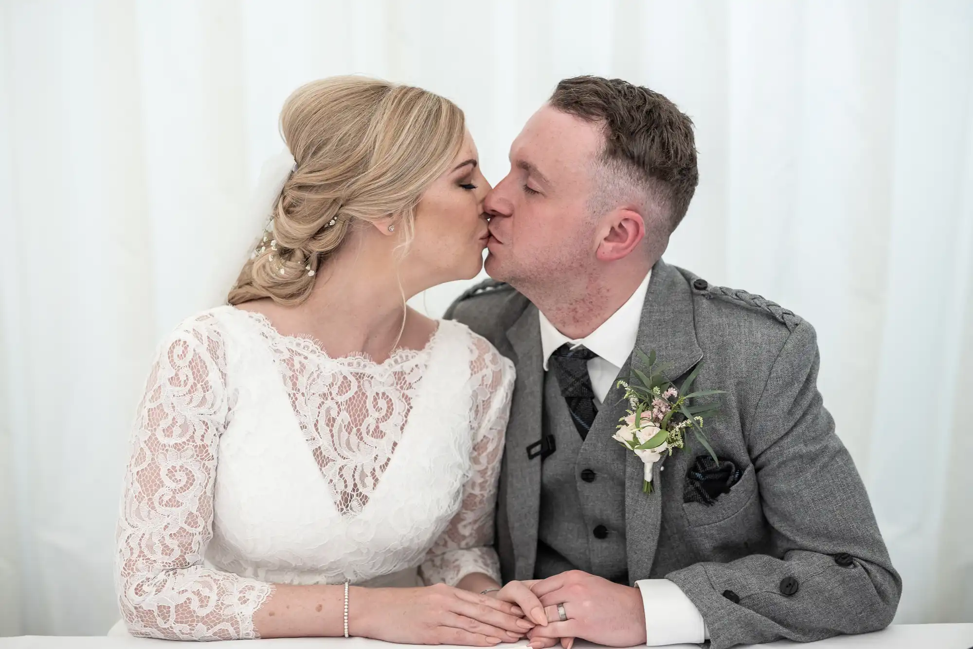 A couple dressed in wedding attire shares a kiss; the bride wears a white lace dress, and the groom wears a gray suit with a boutonniere.