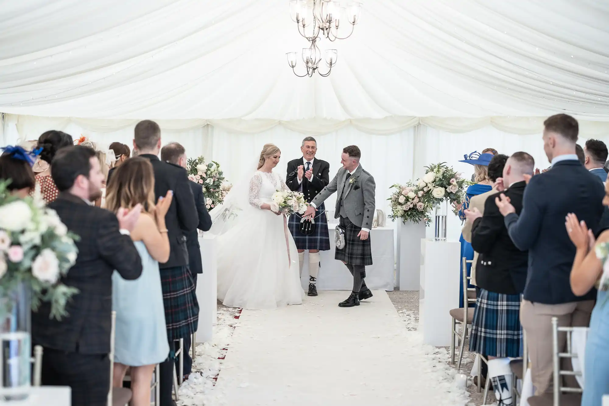 A couple walks down the aisle at their wedding ceremony. Guests are clapping, and a man in a kilt is standing in the background, appearing to officiate the ceremony.