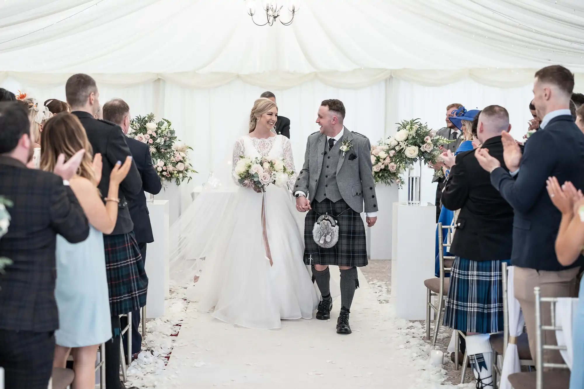 A bride in a white dress and a groom in a kilt walk down the aisle in a tented venue, smiling and holding hands, as guests stand and clap.