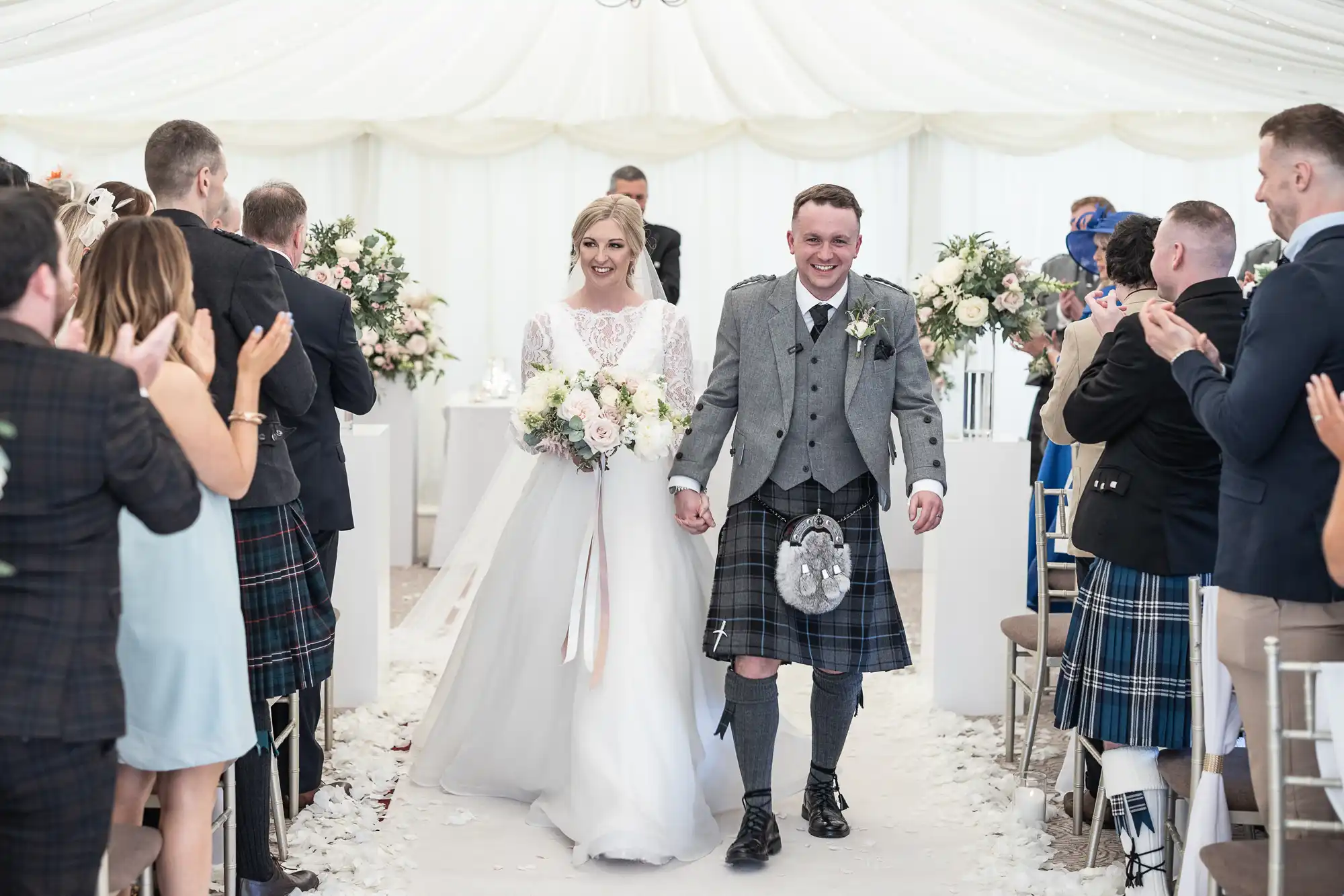 Bride and groom in traditional Scottish attire walk down the aisle, smiling, while guests clap and celebrate in a decorated venue.