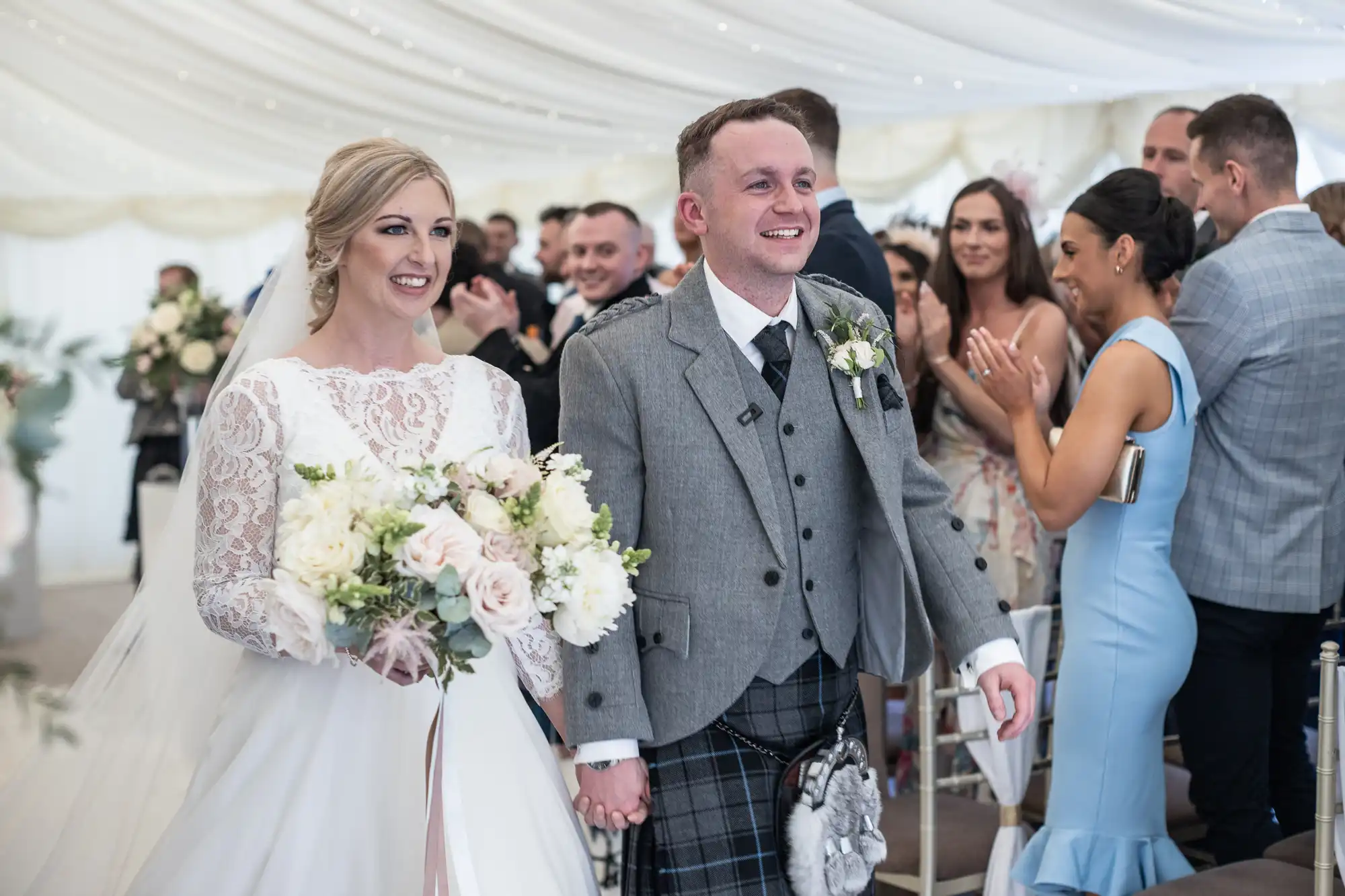 A smiling bride in a white lace dress and a groom in a gray suit walk down the aisle holding hands in a decorated wedding venue while guests applaud.