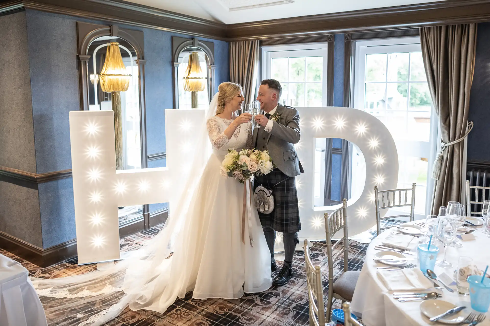 A newly married couple toasts in a decorated room, standing in front of large illuminated letters spelling "H" and "D." The bride is in a white gown, and the groom is in a kilt.