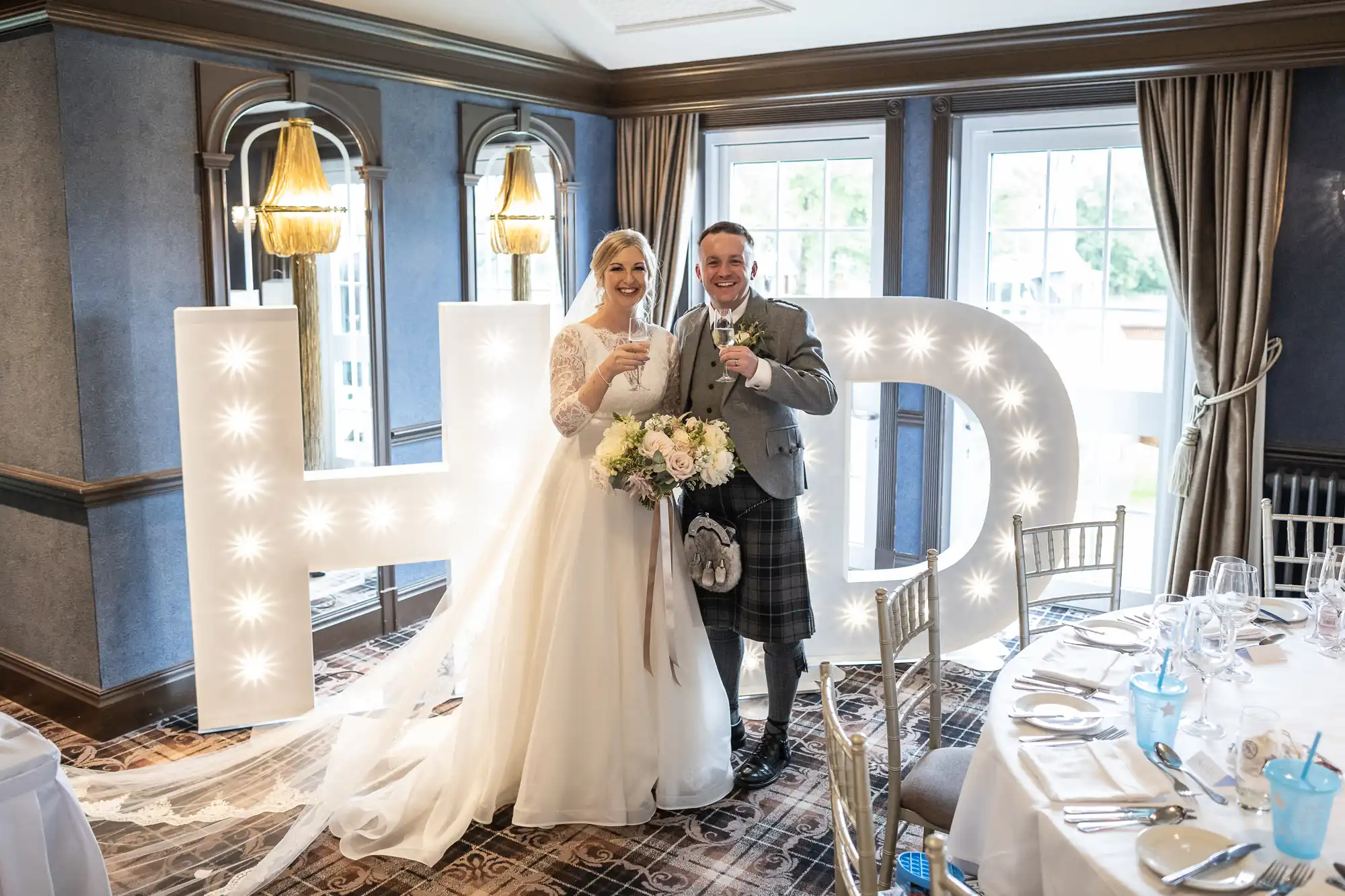 A bride in a white gown and a groom in a kilt pose with drinks in front of large illuminated "H" and "D" letters inside a decorated room.