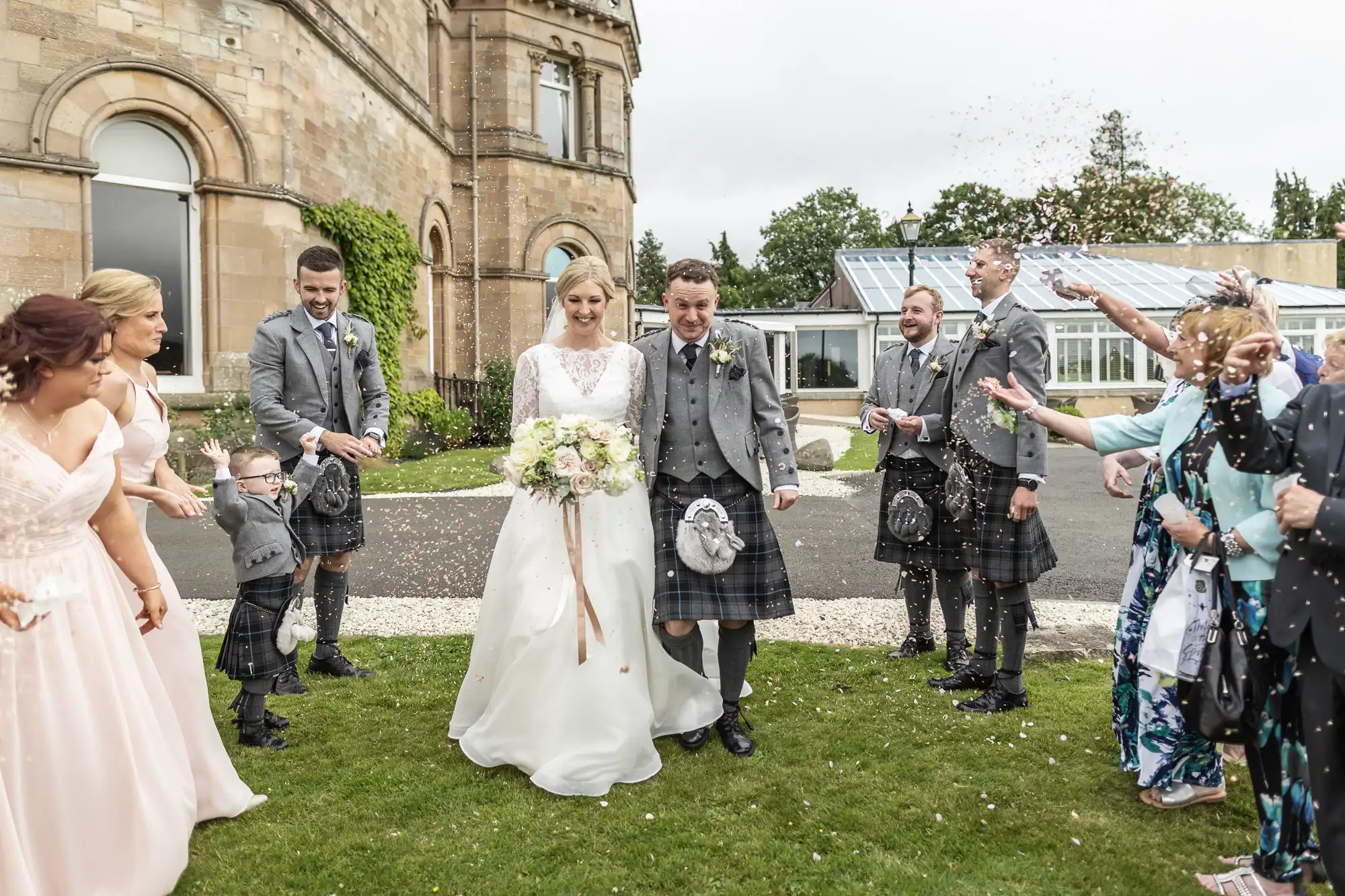 A bride and groom in wedding attire walk outdoors while guests throw confetti. The groom and groomsmen wear kilts, and the bride holds a bouquet. A stone building and greenhouse are in the background.