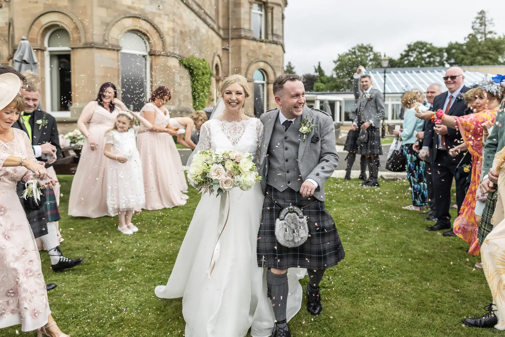 A newlywed couple walks down the aisle outdoors, surrounded by smiling guests in formal attire. The groom wears a kilt, and the bride holds a bouquet of flowers. Confetti is being thrown.