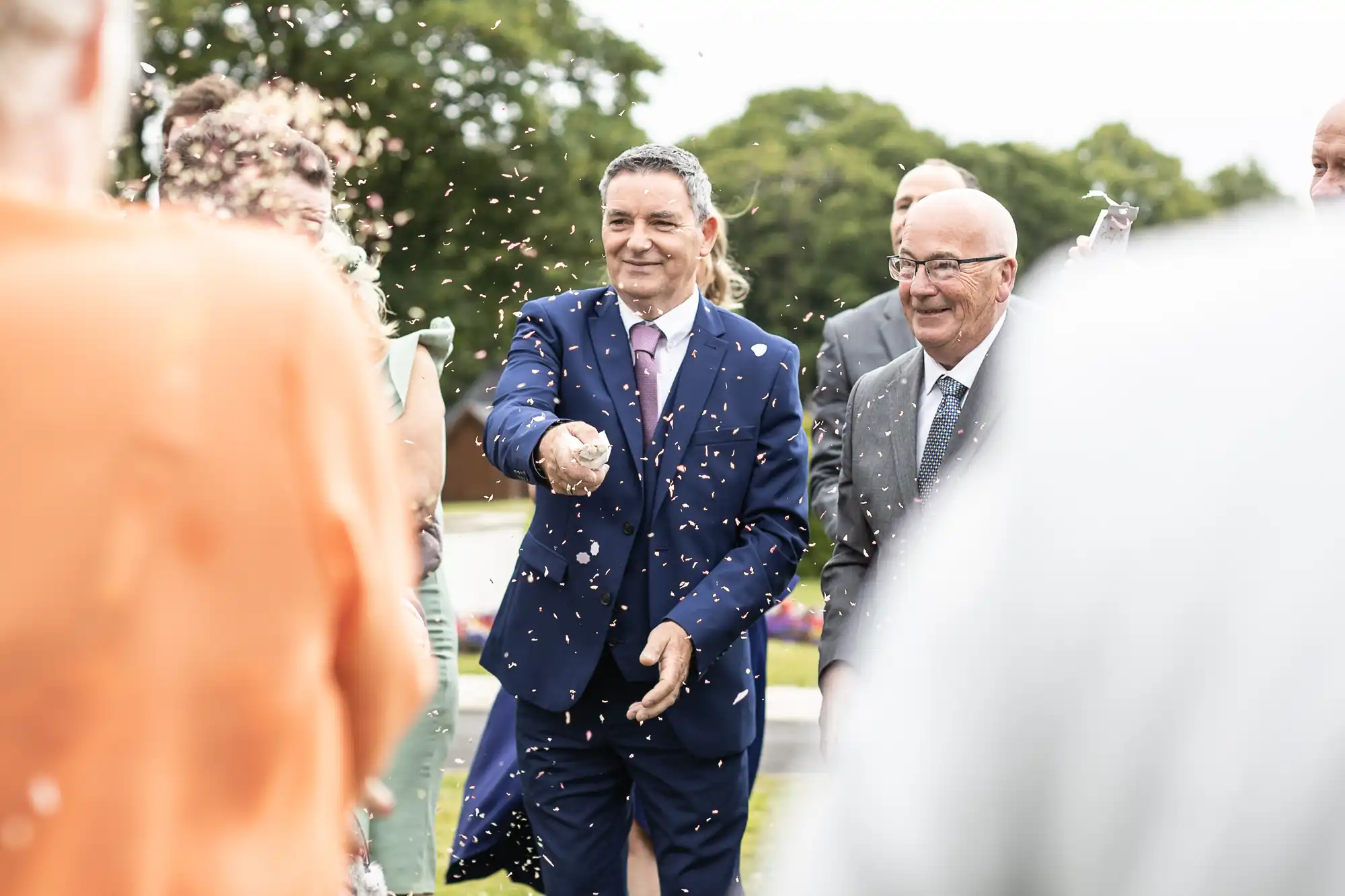 Two men, one in a blue suit and the other in a gray suit, smile and throw confetti outdoors during a celebration. People and greenery are in the background.