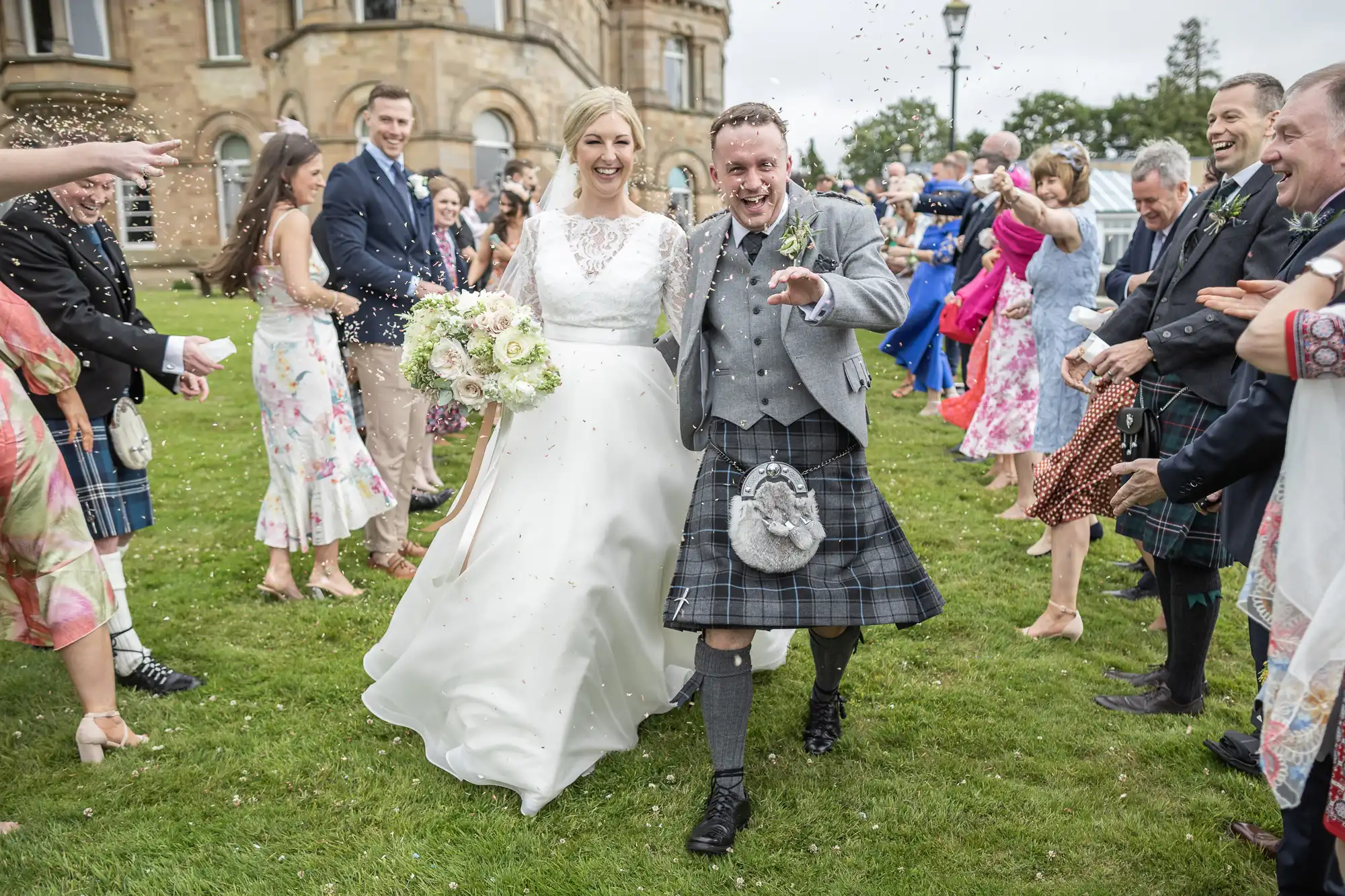 Bride and groom in wedding attire smile and walk through a crowd of guests who are throwing confetti, next to a historic building on a grassy lawn.