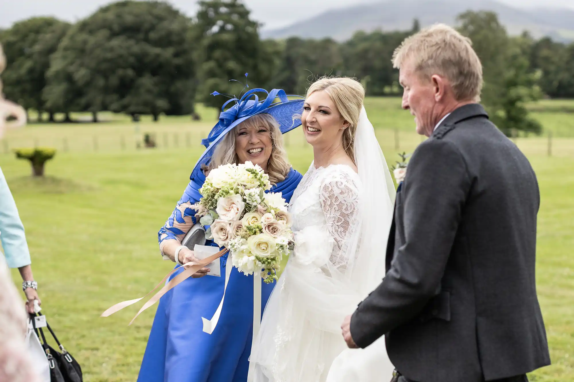 Bride in a white dress holds a bouquet, smiling with a woman in a blue dress and hat, while a man looks on. They are outside with greenery in the background.