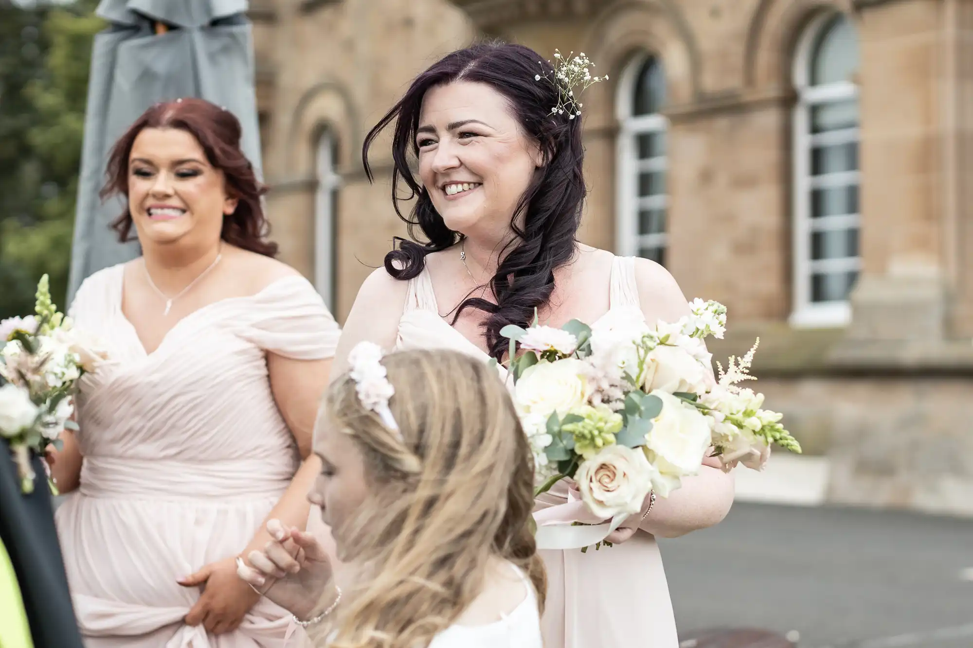 Women in light pink dresses holding floral bouquets, standing outdoors in front of a stone building.