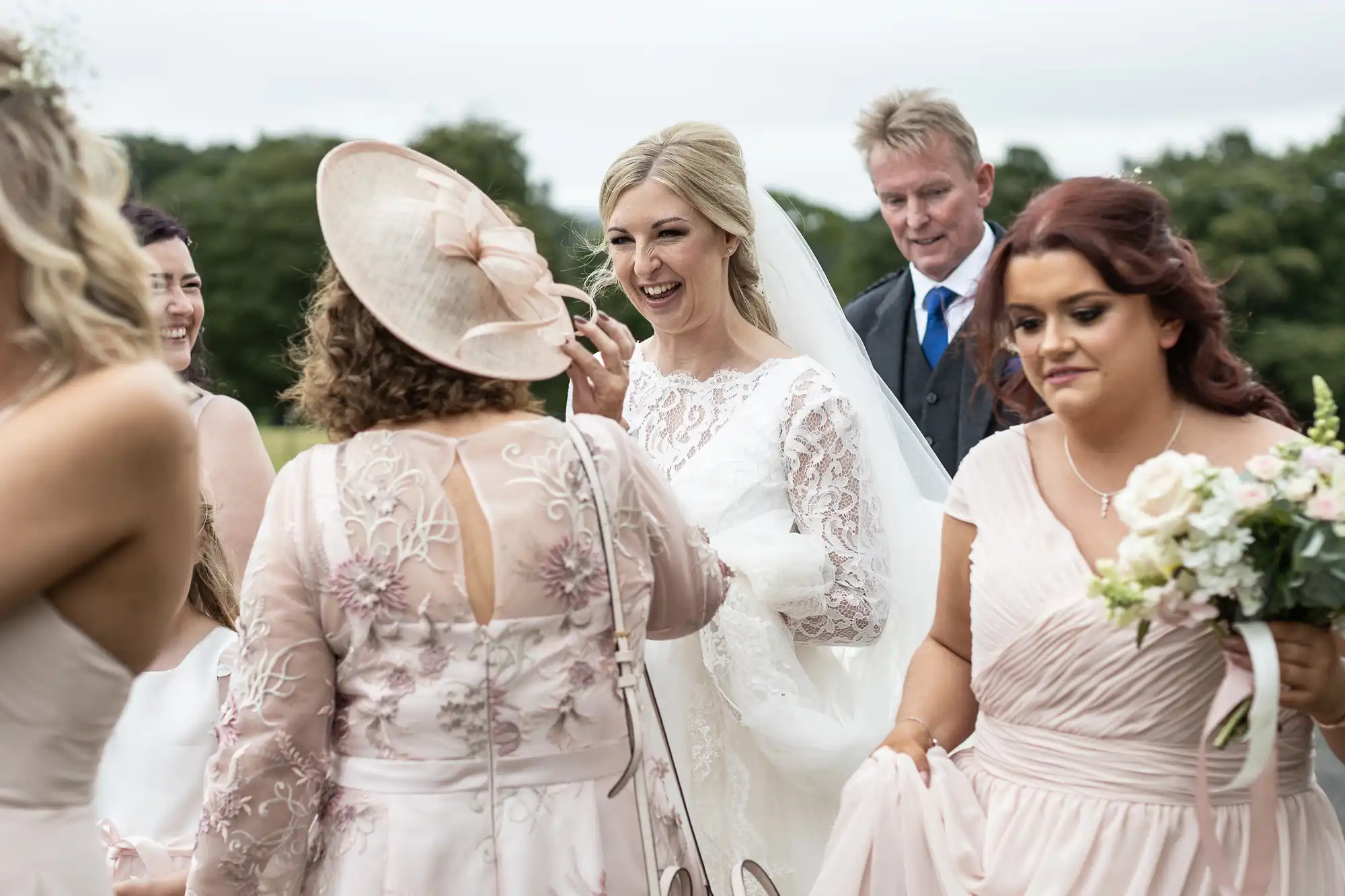 A bride in a white gown smiles while standing among bridesmaids in light pink dresses and a few guests outdoors. One bridesmaid holds a bouquet, and a man in a suit stands in the background.