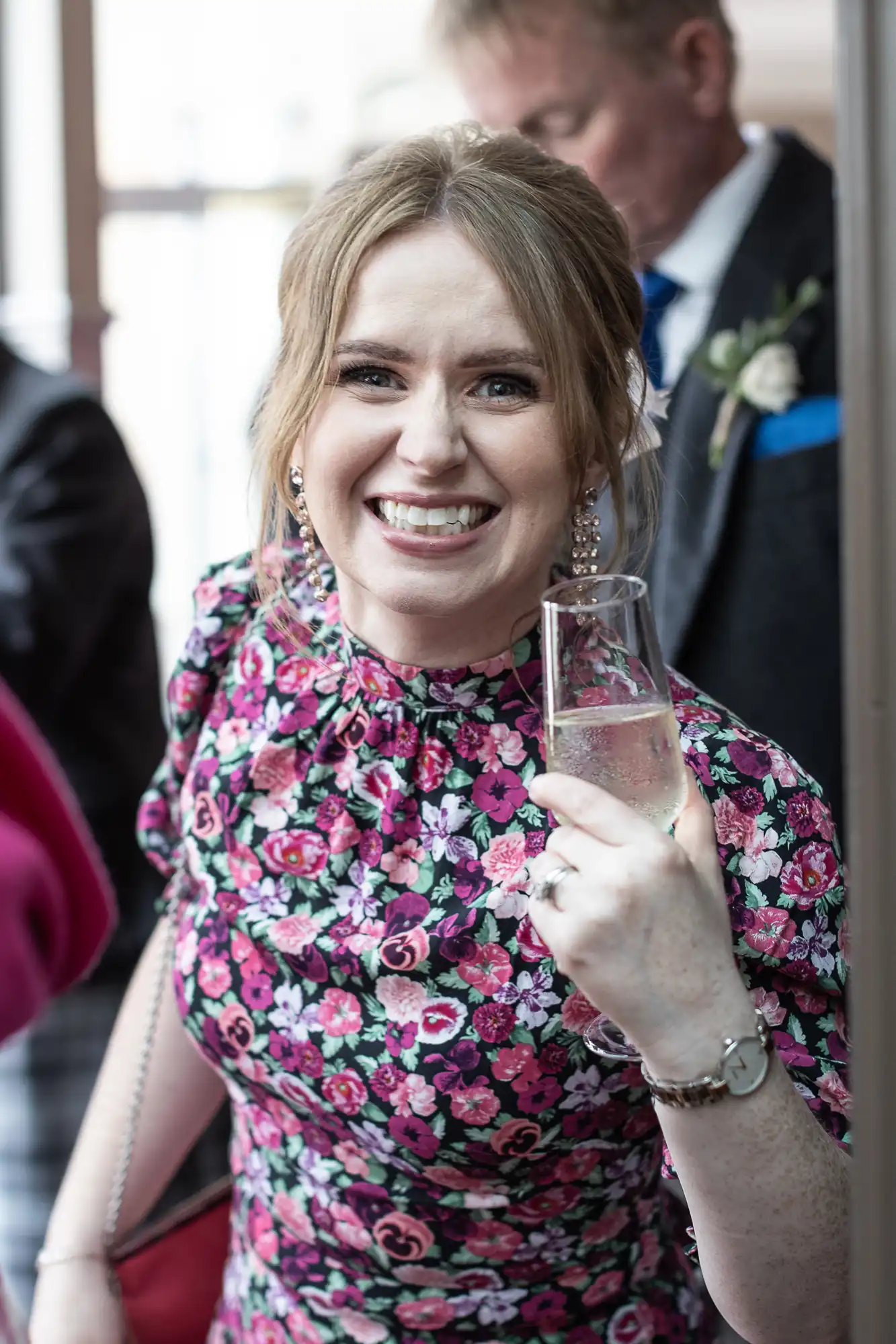 A woman in a floral dress smiles while holding a glass of champagne. Other people can be seen in the background.