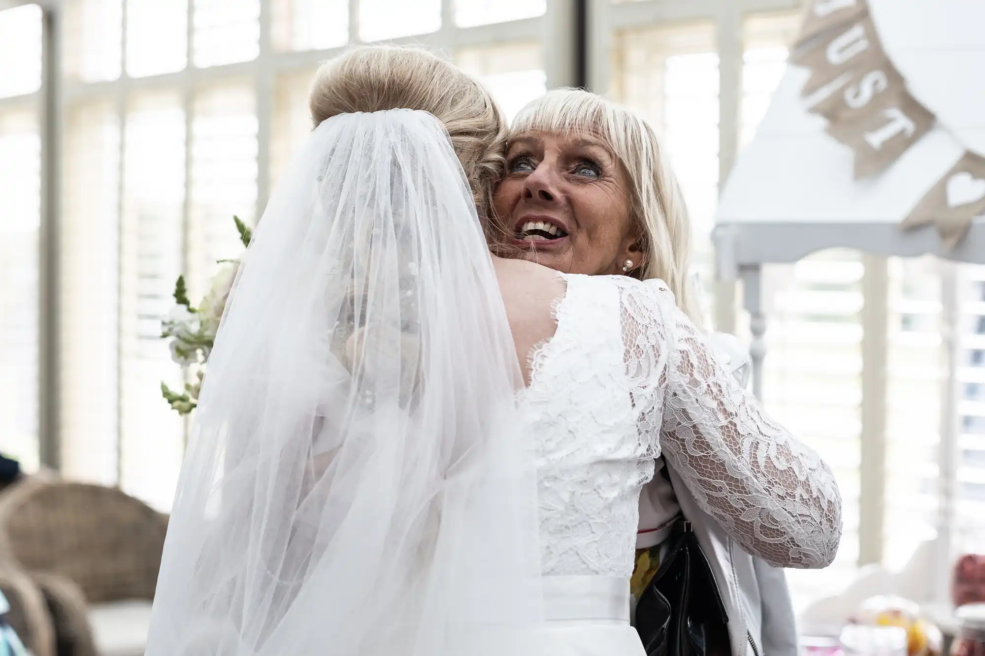 A bride in a white dress and veil embraces an older woman in a white outfit at an indoor event.