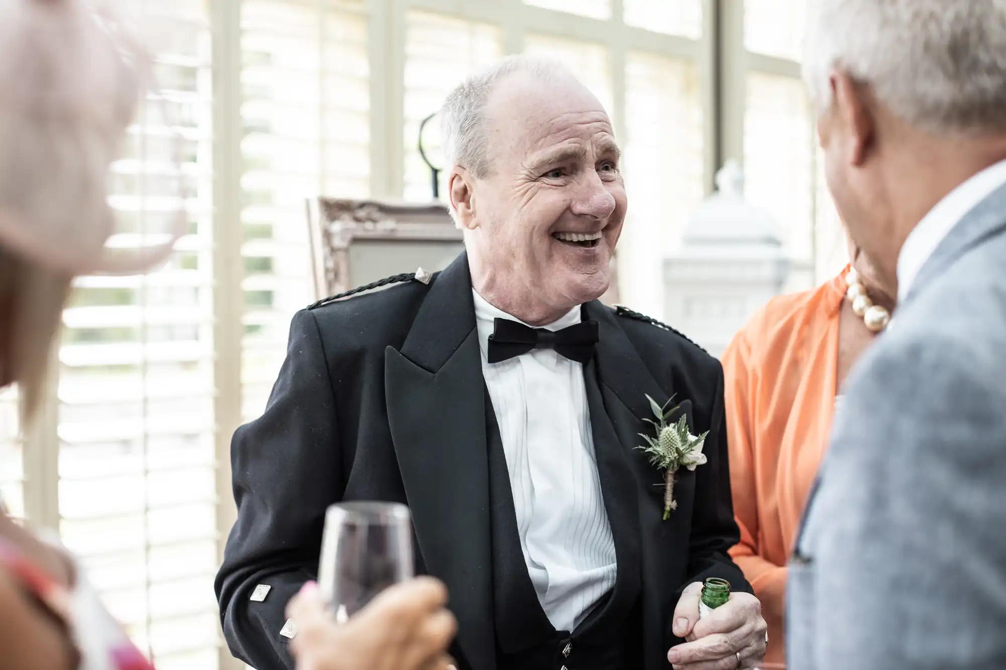 An elderly man in a black tuxedo and bow tie smiles while holding a green bottle and talking to three other people in formal attire.