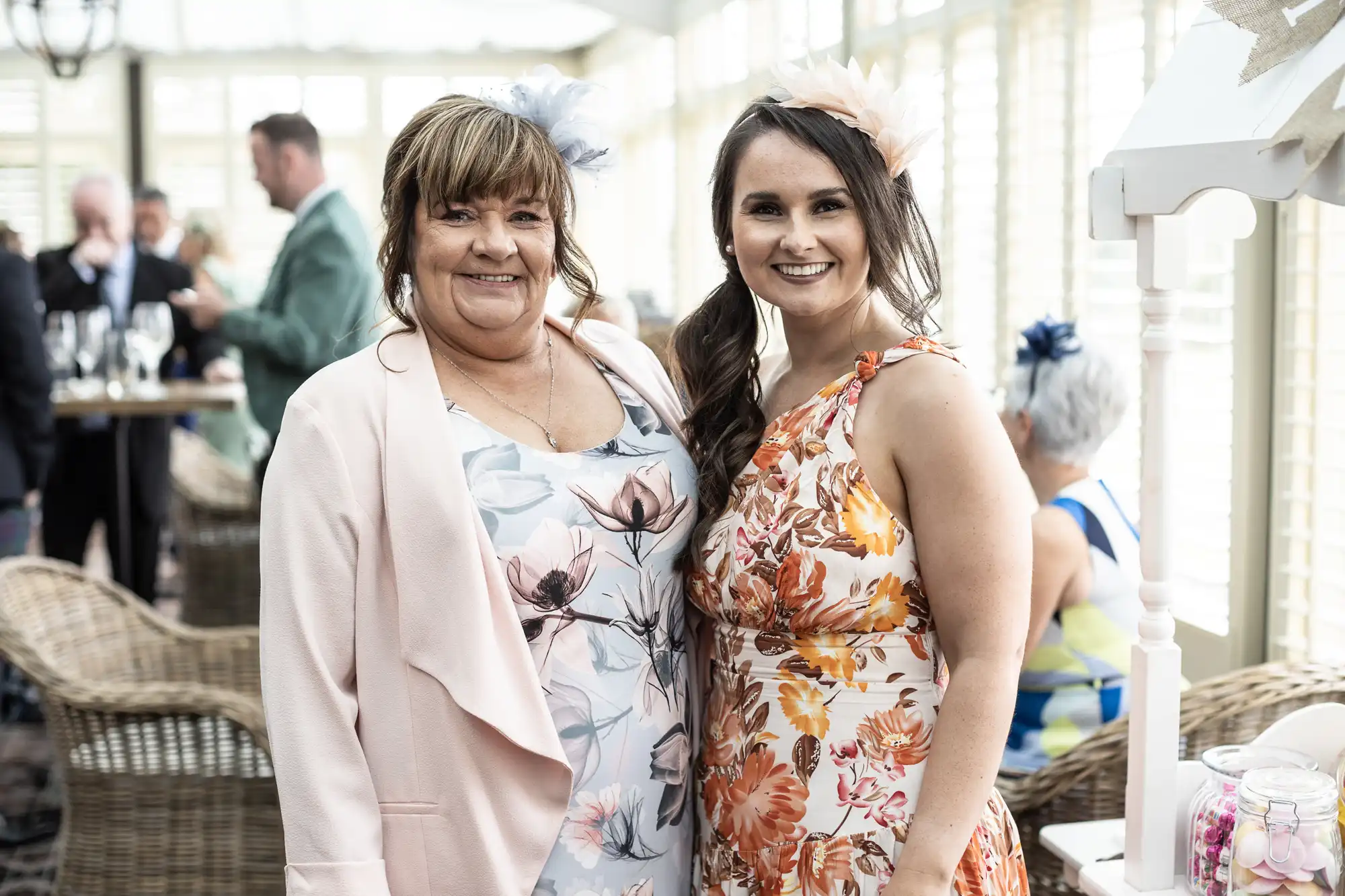 Two women smiling and posing together at a social event, wearing floral dresses and fascinators. Other attendees are in the background, sitting and standing in a wicker-furnished room.