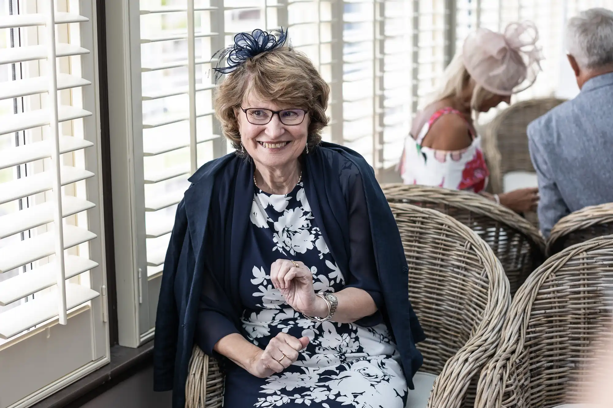 An older woman with glasses, wearing a navy blue floral dress and matching hat, sits on a wicker chair inside a room with large windows. Two other people are seated behind her.