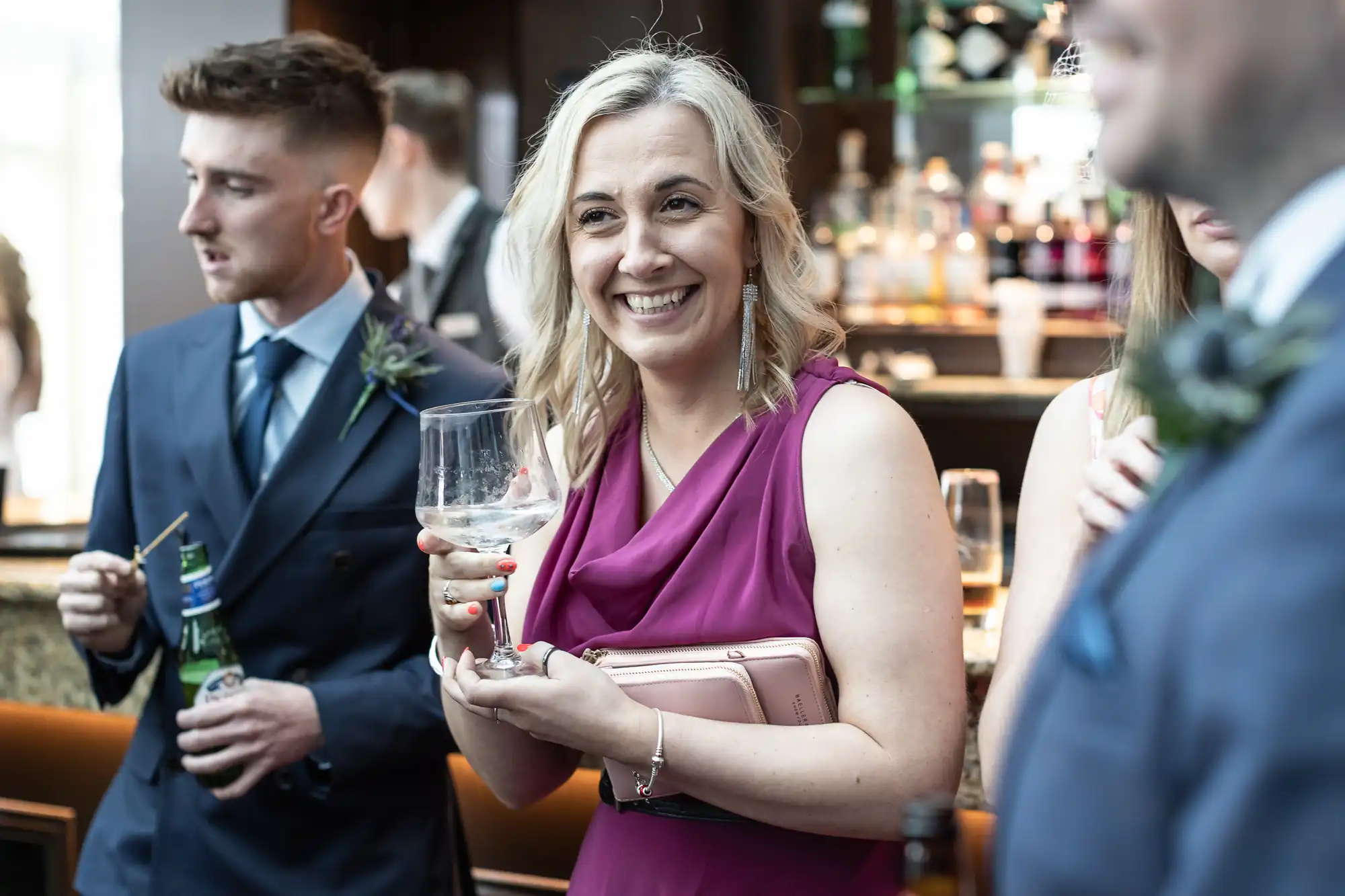 A woman in a magenta dress smiles while holding a wine glass at a social event with other people in the background.