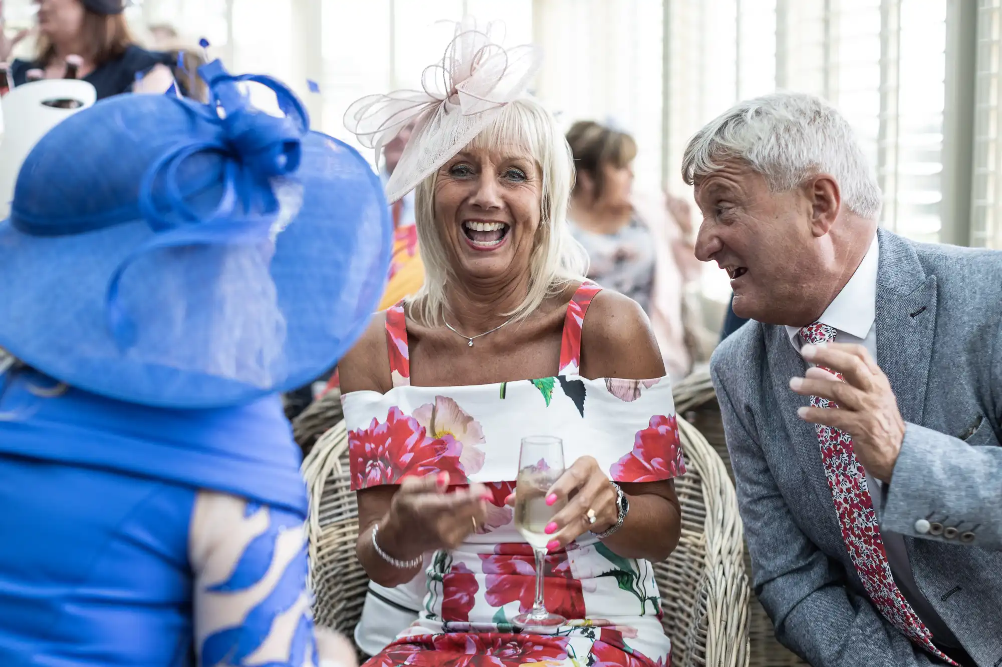 A woman in a floral dress and fascinator is laughing while holding a drink, seated next to a man in a grey suit and tie who is speaking. Both are facing a person in a blue outfit with a large hat.