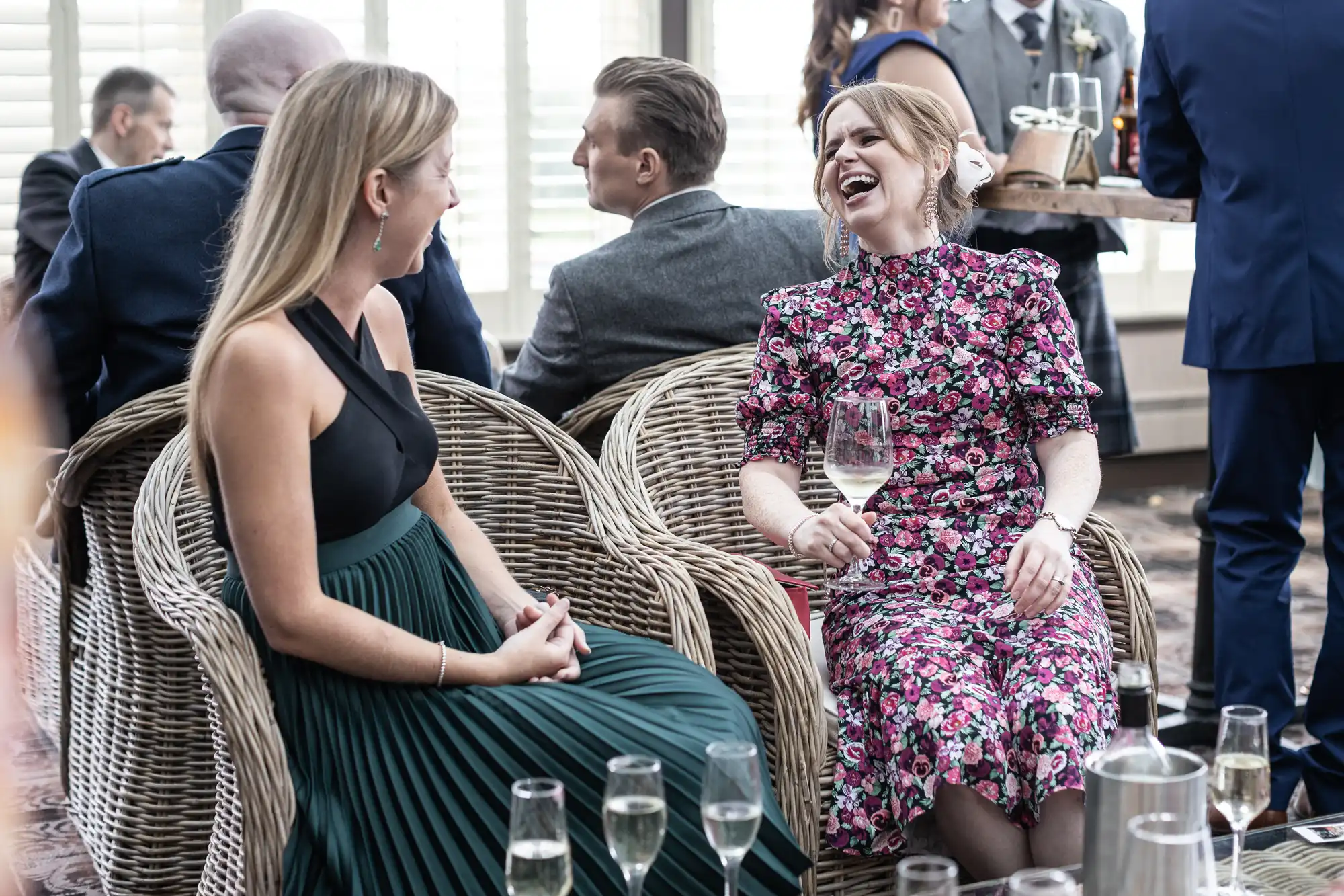 Two women sit in wicker chairs, laughing and chatting at a social event. One wears a black and green dress, and the other wears a floral-patterned dress while holding a glass of wine.