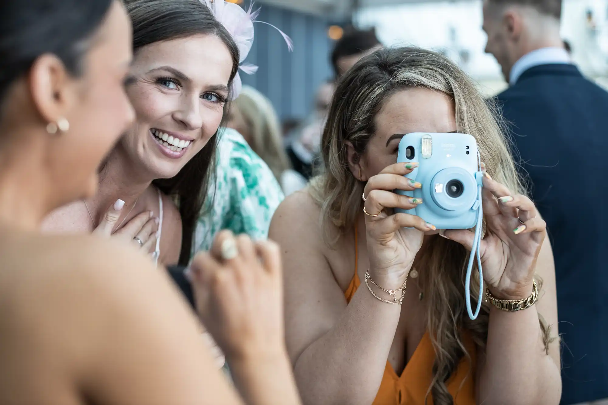 Two women are smiling at the camera, while another woman in an orange dress uses a light blue instant camera to take a photo. People are visible in the background at what appears to be an outdoor event.